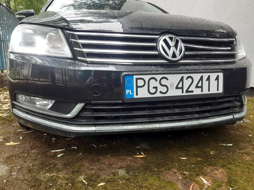 Zbliżenie na przód czarnego samochodu marki Volkswagen. Widoczny jest grill z logo marki, reflektory oraz polska tablica rejestracyjna o numerze PGS 42411. Samochód stoi na podłożu z mchu i ziemi, na którym leżą pojedyncze liście.
