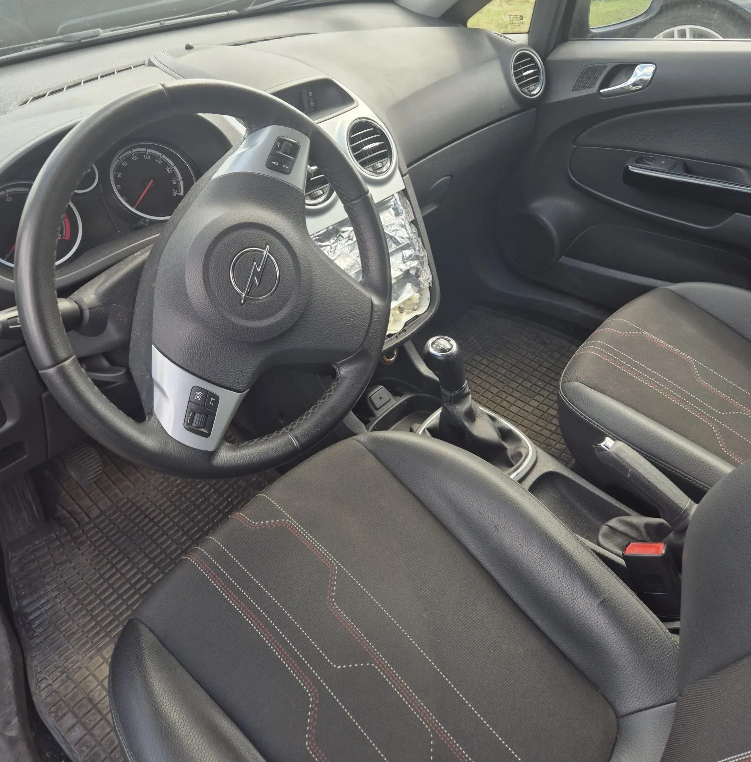 Wnętrze samochodu marki Opel, widok z perspektywy kierowcy. Widoczna jest czarna kierownica z logo producenta, deska rozdzielcza z licznikiem, dwa przednie fotele z czarnej tkaniny z czerwonymi przeszyciami oraz lewarek zmiany biegów. Część konsoli środkowej jest owinięta srebrną folią ochronną. Na podłodze znajdują się gumowe dywaniki z wypustkami.