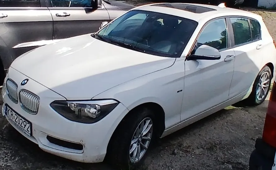Biały samochód marki BMW w nadwoziu hatchback, zaparkowany na utwardzonym placu. Widoczny jest przód i prawy bok pojazdu, tablica rejestracyjna o numerze KR 2U292 oraz szyberdach. W tle widoczny fragment innego, ciemnego samochodu.