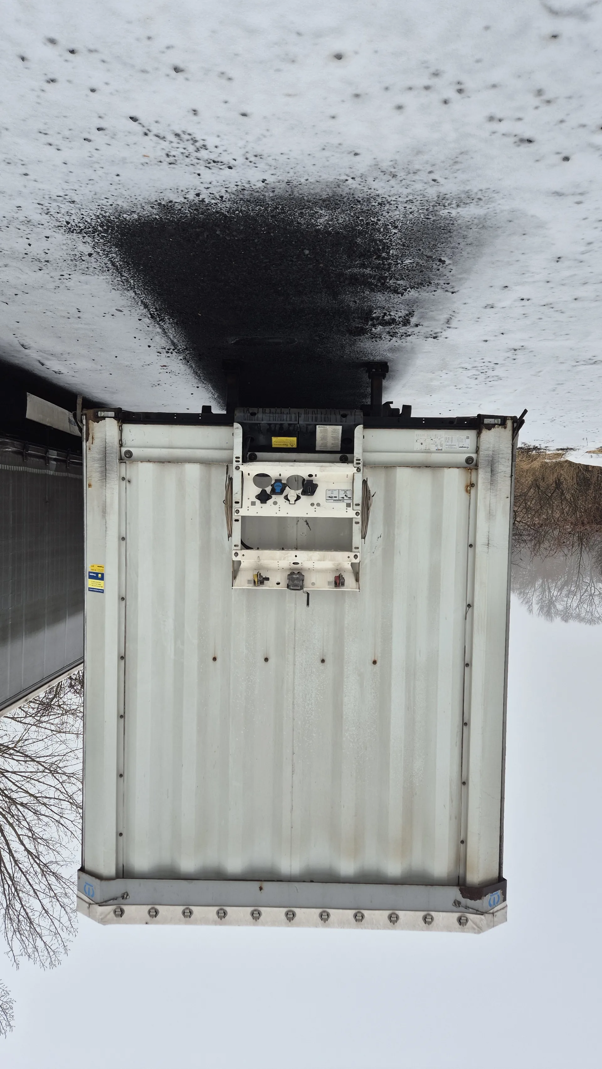 Biały przyczepa chłodnicza z widocznymi elementami mechanicznymi na tylnej ścianie. Na przyczepie znajdują się małe, nieczytelne etykiety i naklejki. Tło stanowi śnieżna powierzchnia z ciemnymi plamami, nagie drzewa oraz pochmurne niebo. Przyczepa jest ustawiona pionowo, co sugeruje nieprawidłową orientację obrazu.