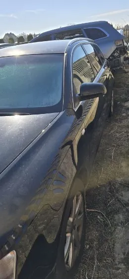 Ciemny samochód osobowy z widocznymi uszkodzeniami na boku, zaparkowany na suchym, trawiastym terenie. Za nim znajduje się uszkodzony pojazd typu SUV. Tło stanowi niebo oraz odległe drzewa. Słoneczne oświetlenie rzuca cienie na powierzchnię samochodów.