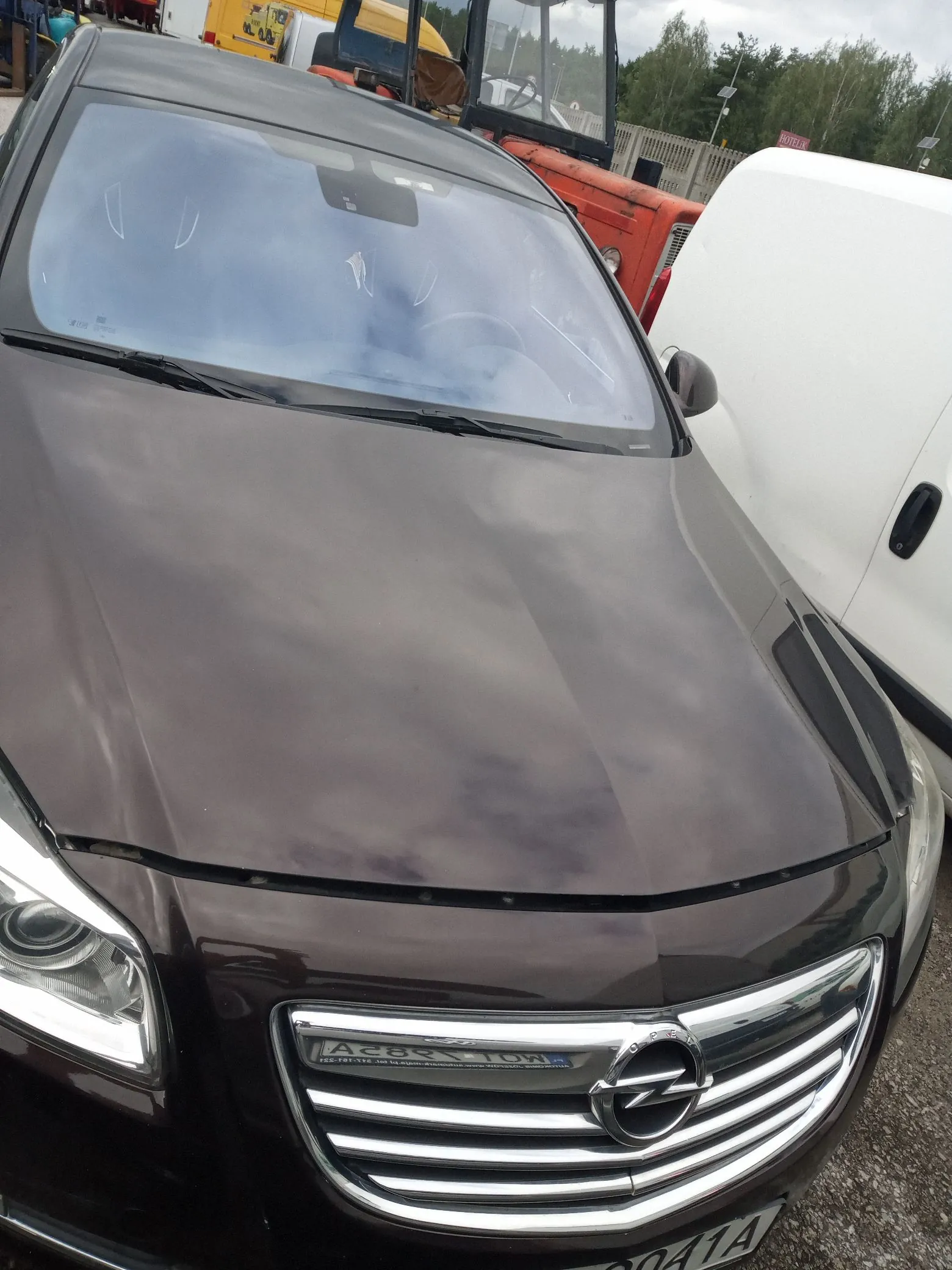 Zbliżenie na przód ciemnobrązowego samochodu marki Opel. Widoczna jest maska, przednia szyba oraz grill z logo producenta. Na grillu znajduje się naklejka z napisem 'A285 TOWN'. Obok samochodu stoi biały pojazd, a w tle widoczny jest czerwony ciągnik rolniczy oraz drzewa.