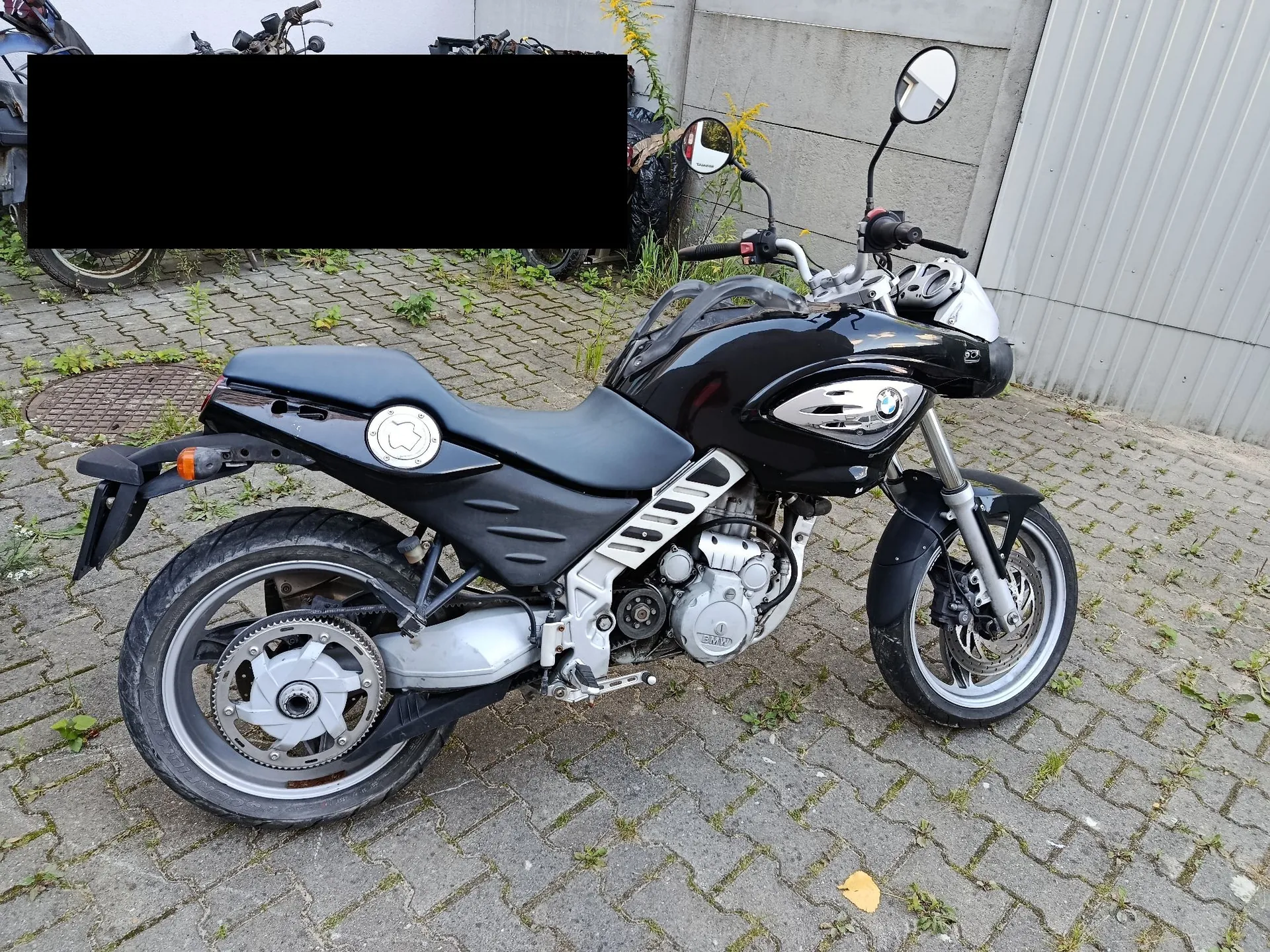 Czarny motocykl marki BMW zaparkowany na kostce brukowej przed szarym murem i metalową bramą. Motocykl jest ustawiony bokiem, widoczny jest silnik, koła, siedzenie oraz lusterka.