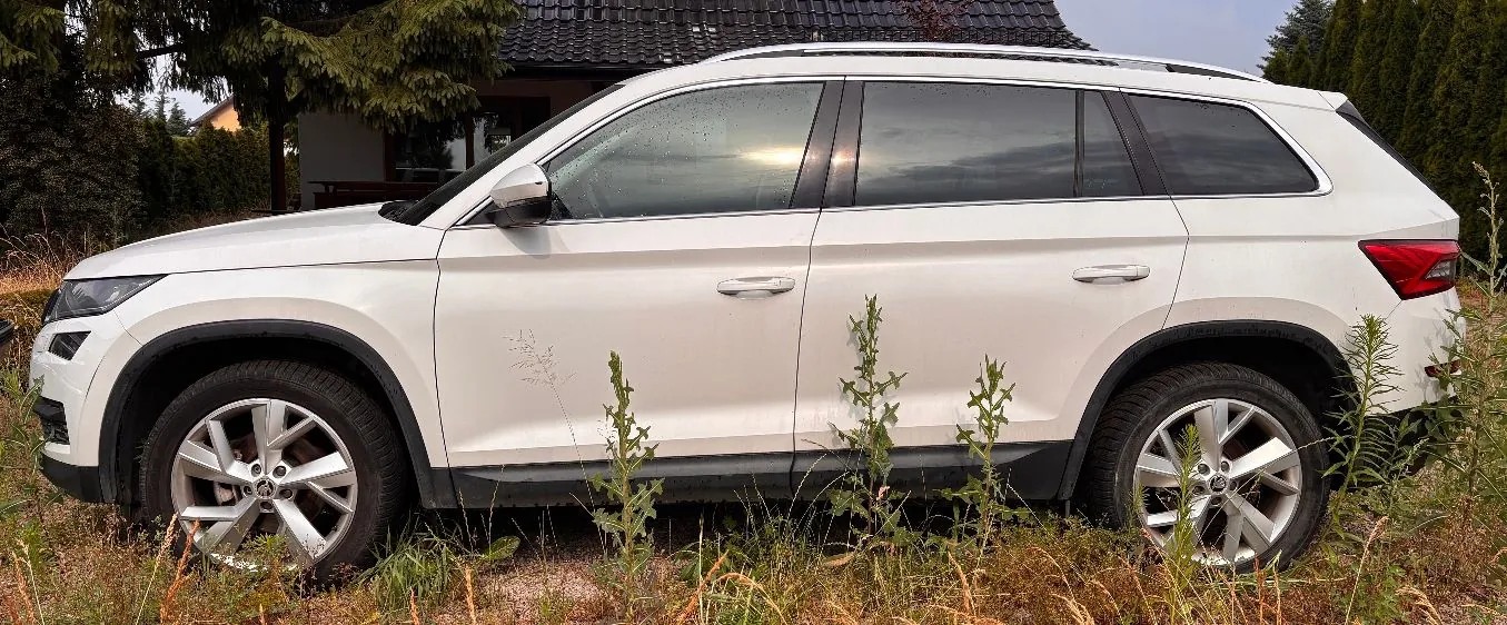 Biały samochód typu SUV zaparkowany na trawie przed domem z ciemnym dachem. Widok z profilu na prawą stronę pojazdu.