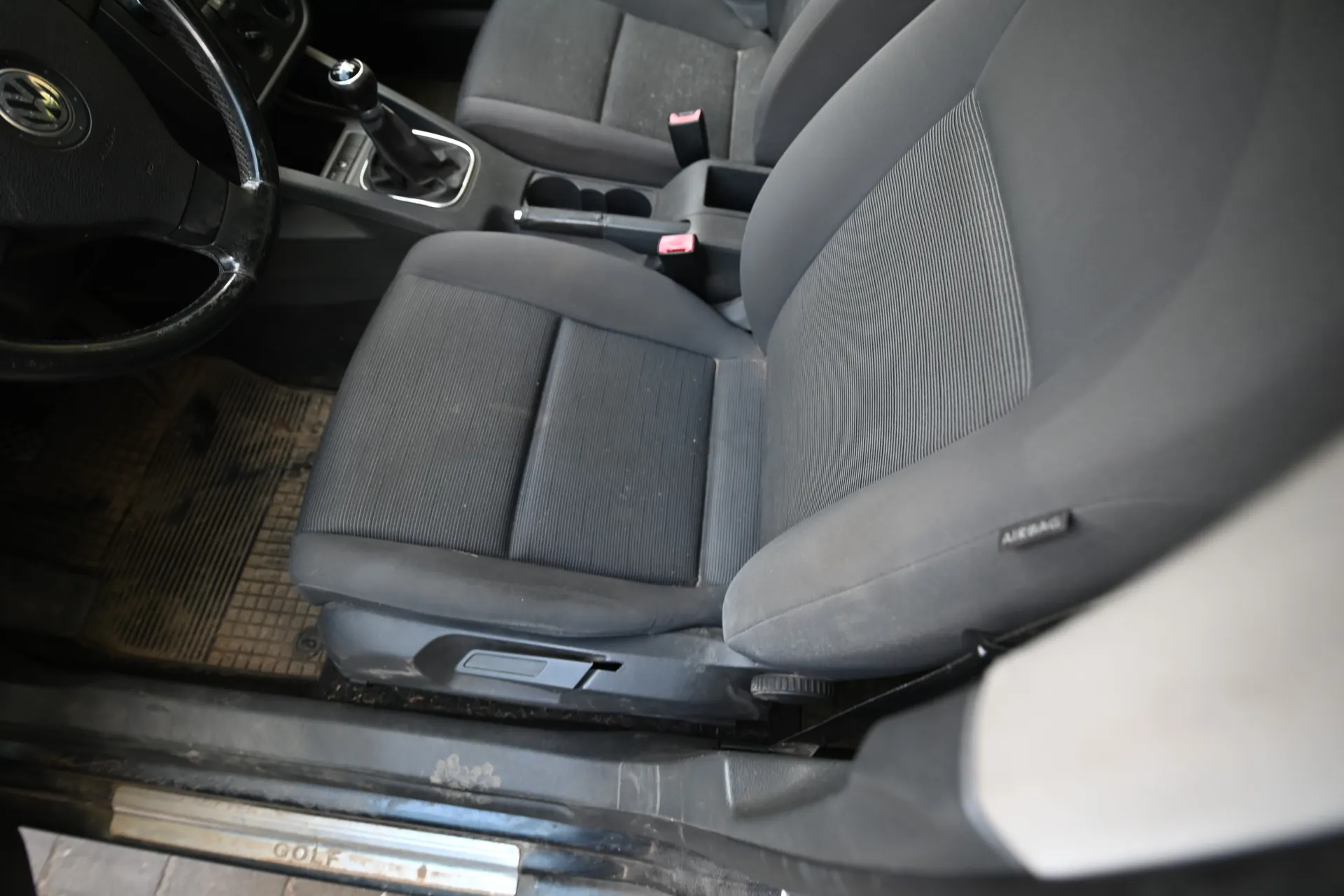 Wnętrze samochodu marki Volkswagen. Widoczna kierownica z logo VW po lewej stronie, szare tkaninowe siedzenia, dźwignia zmiany biegów w centralnej konsoli, brudne dywaniki podłogowe oraz napis "GOLF" na progu drzwi. Na siedzeniu pasażera widoczny napis "AIRBAG". W centralnej konsoli znajdują się uchwyty na napoje i dźwignia hamulca ręcznego z czerwonym przyciskiem.