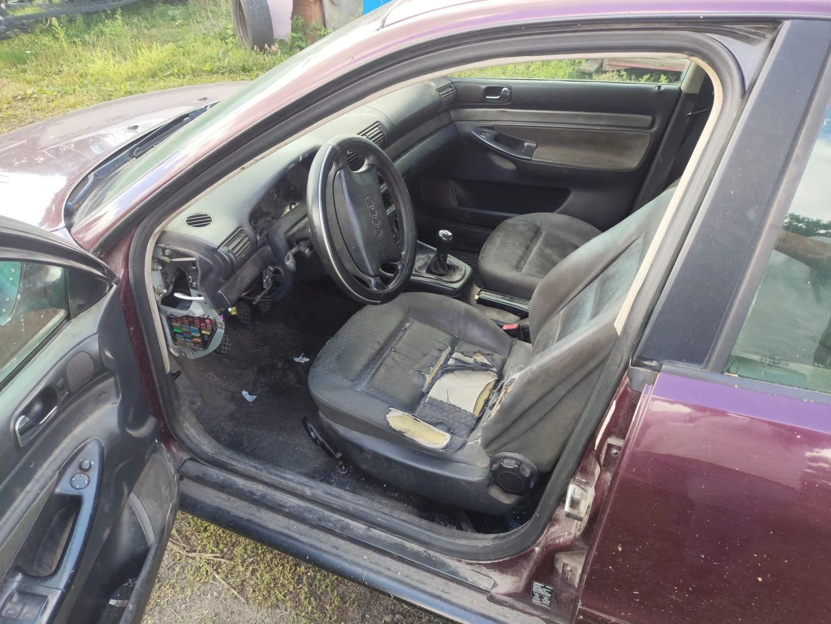 Wnętrze bordowego samochodu z otwartymi drzwiami od strony kierowcy. Widoczna jest kierownica z logo Audi, deska rozdzielcza z brakującymi elementami oraz przedni fotel kierowcy z wyraźnymi przetarciami i dziurami w materiale tapicerki. Wewnątrz pojazdu widoczne są zanieczyszczenia.