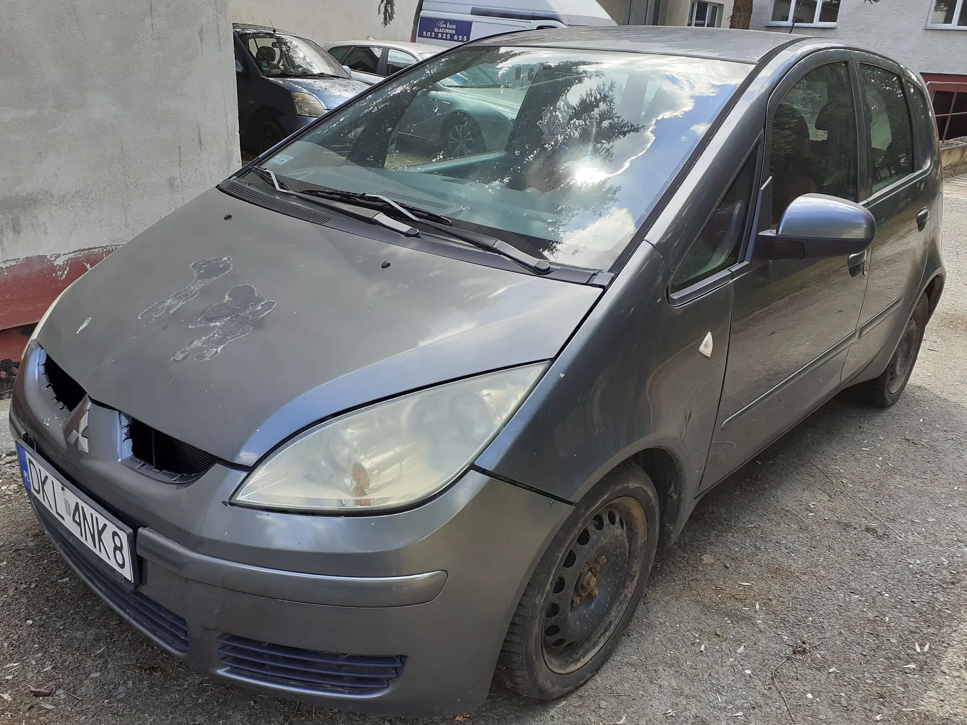 Srebrnoszary samochód osobowy marki Mitsubishi zaparkowany na utwardzonym podłożu obok jasnej ściany budynku. Pojazd posiada widoczne uszkodzenia lakieru na masce oraz zmatowione reflektory. Na przednim zderzaku znajduje się tablica rejestracyjna o numerze DKL 4NK8.