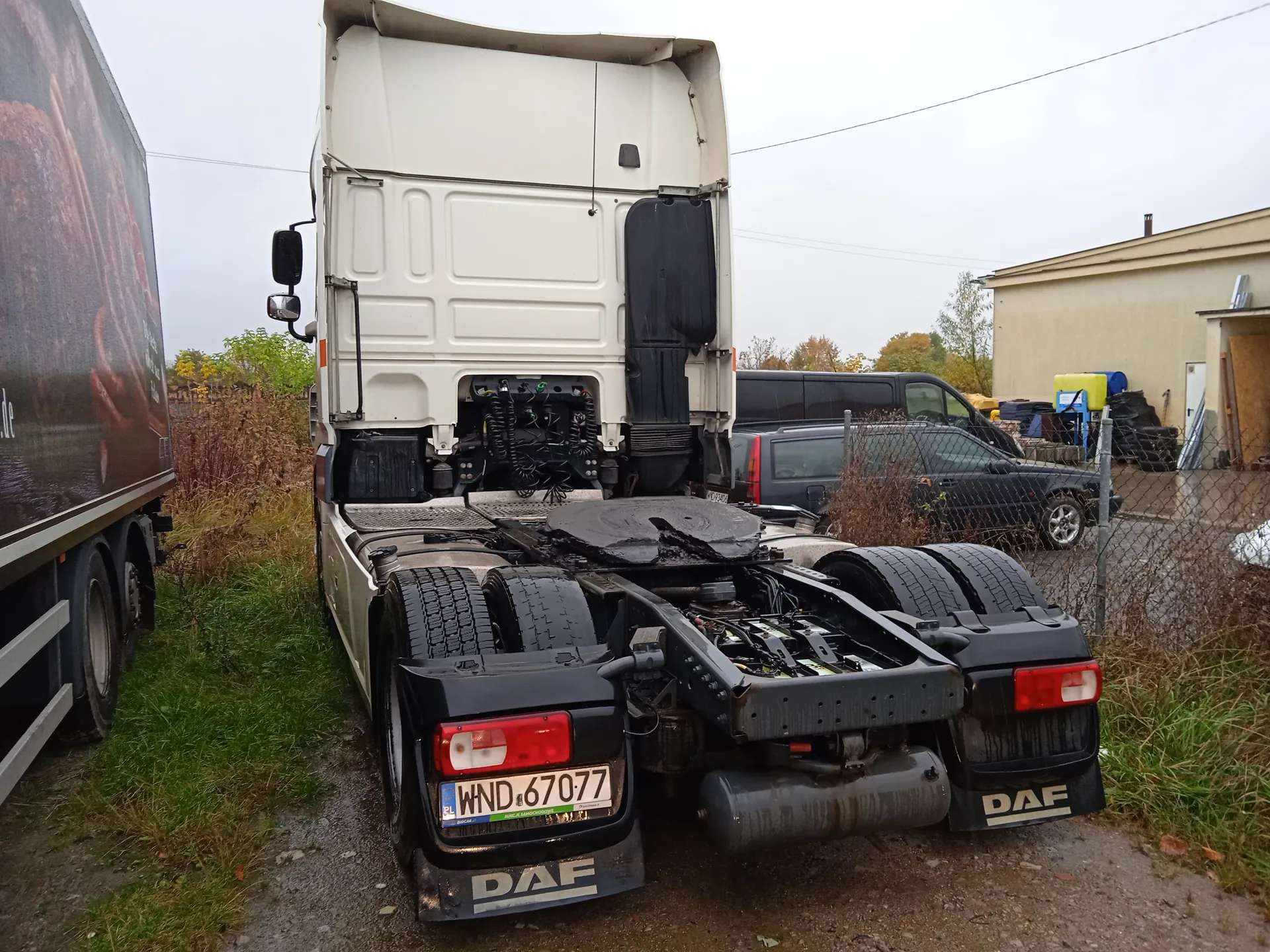 Biały ciągnik siodłowy DAF bez przyczepy, widok z tyłu, tablica rejestracyjna WND 67077. Odsłonięte elementy techniczne, cztery opony. Po lewej ciężarówka z częściowo widoczną reklamą. W tle ogrodzenie, czarny samochód, budynek i trawa na żwirowej nawierzchni.