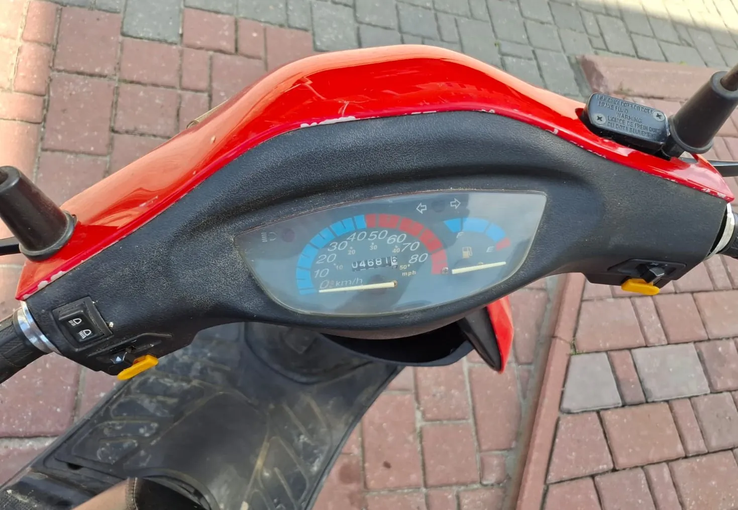 Zbliżenie na kokpit czerwonego skutera. Widoczny jest analogowy prędkościomierz z podziałką w km/h i mph, wskaźnik poziomu paliwa oraz licznik kilometrów wskazujący wartość 04881. Po bokach znajdują się czarne manetki z przełącznikami. Skuter stoi na kostce brukowej.