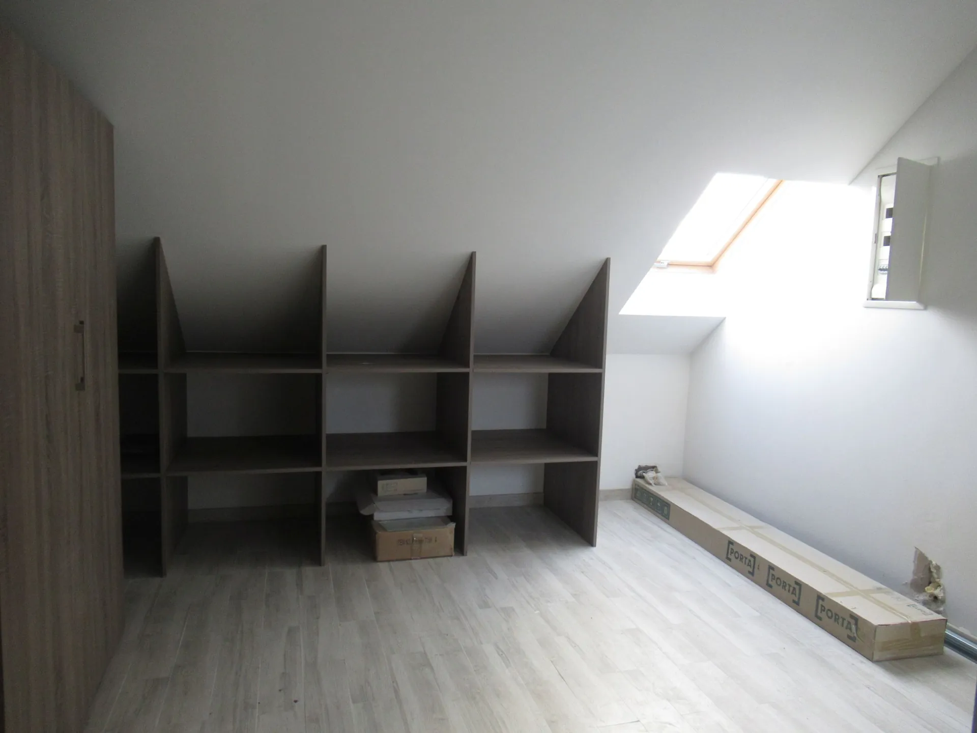 Wnętrze jasnego pomieszczenia z białymi ścianami i jasną drewnianą podłogą. Pod skosem dachu znajduje się wbudowany regał z szarymi półkami, podzielony na kilka sekcji. Na jednej z dolnych półek leżą białe pudełka i karton. Po prawej stronie znajduje się okno dachowe, przez które wpada jasne światło. Na podłodze, przy prawej ścianie, leży długa, wąska paczka z napisem 'PORTA' powtórzonym kilka razy. Po lewej stronie widoczny jest fragment szafy w kolorze drewna.