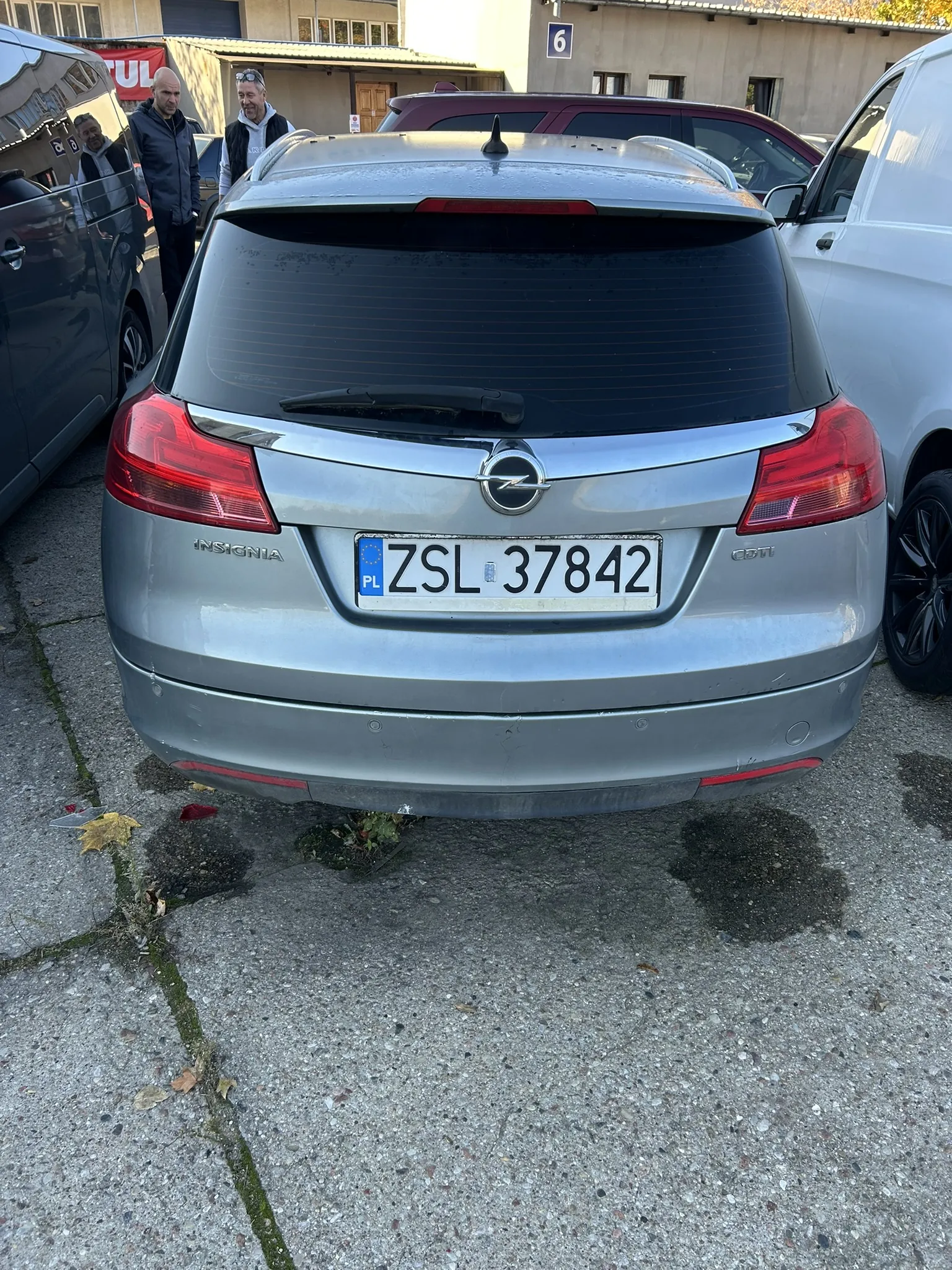 Tył srebrnego samochodu Opel Insignia z tablicą rejestracyjną ZSL 37842 (PL). Po lewej stronie ciemny van, po prawej biały van. W tle budynki, w tym jeden z tablicą numeru 6 oraz dwaj mężczyźni stojący blisko ciemnego vana. Na asfalcie widoczne plamy i liście.
