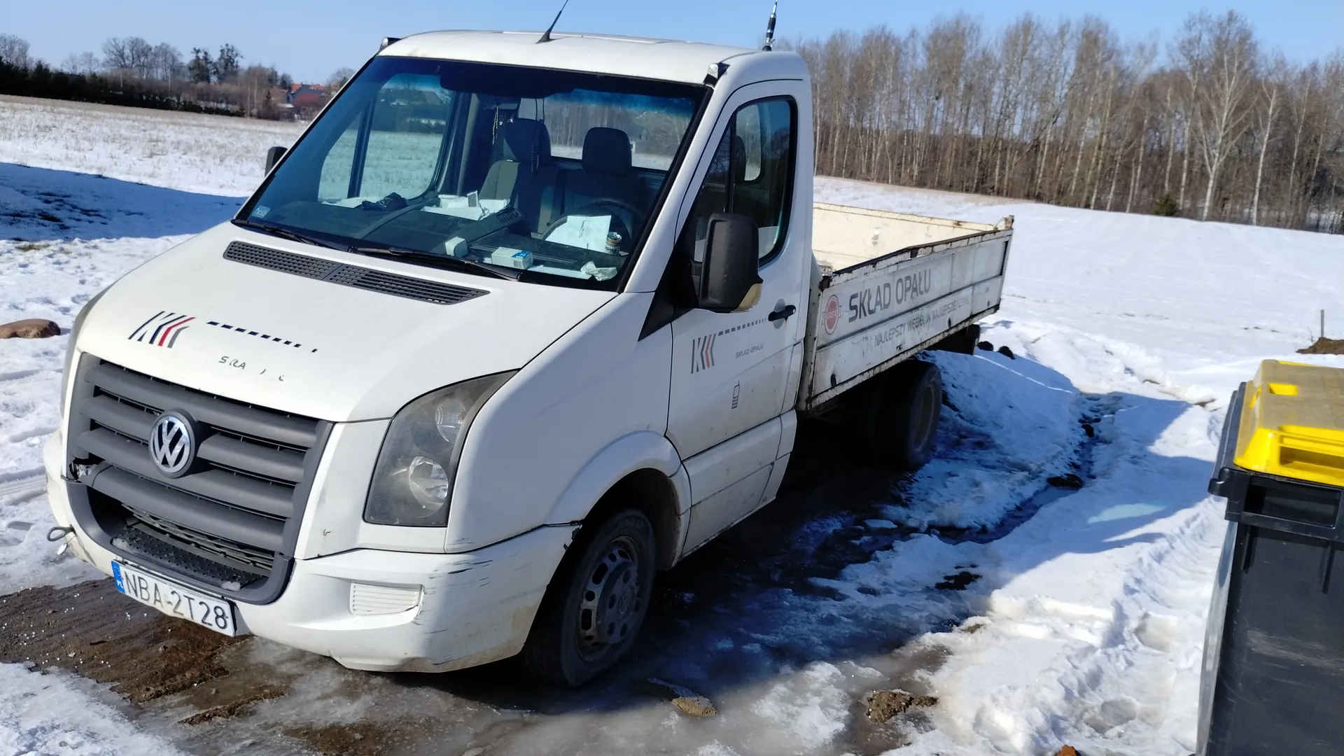 Biały samochód dostawczy marki Volkswagen z otwartym przyczepem, zaparkowany na śnieżnej powierzchni. Na boku przyczepy widoczny napis 'SKŁAD OPAŁU' oraz logo 'KKK' na masce i drzwiach. Tablica rejestracyjna 'NBA-2T28'. W tle pola pokryte śniegiem, drzewa bez liści i jasne niebo. Po prawej stronie obrazu czarno-żółty pojemnik. Wewnątrz kabiny widoczne dokumenty i przedmioty na desce rozdzielczej.