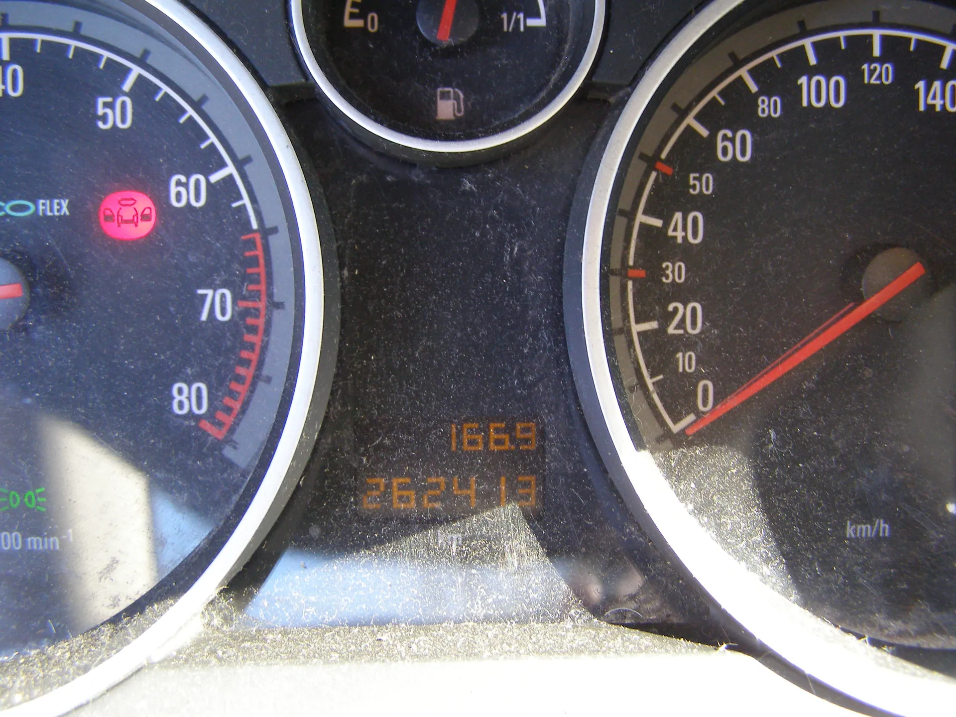 Zbliżenie na deski rozdzielcze samochodu. Po lewej stronie wskaźnik obrotomieru z zakresem do 80, czerwonym symbolem ostrzegawczym przedstawiającym samochód z otwartymi drzwiami oraz napisem FLEX. W centrum wskaźnik paliwa z oznaczeniem E0 i ikoną zbiornika paliwa. Po prawej stronie wskaźnik prędkości z zakresem do 140 km/h, czerwoną igłą wskazującą 0 km/h i napisem km/h. W dolnej części wyświetlacze cyfrowe z wartościami 1669 i 262413. Na desce rozdzielczej widoczny jest kurz. W lewym dolnym rogu zielony napis 00 min-1.