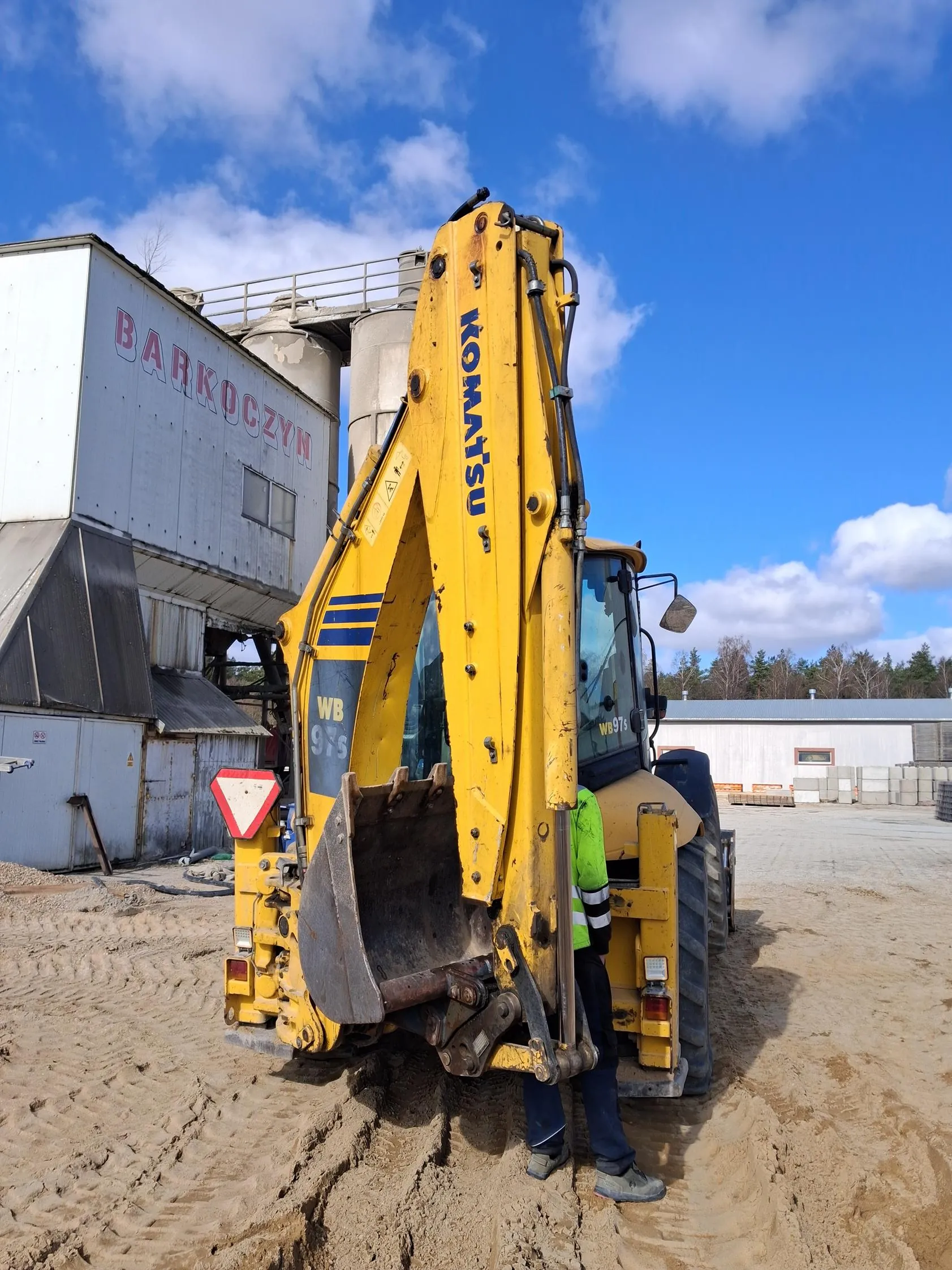 Żółty koparko-ładowarka marki Komatsu model WB 97S na terenie budowlanym. Na maszynie widoczny napis KOMATSU oraz oznaczenie WB 97S. Obok maszyny stoi osoba w zielonej kamizelce odblaskowej i ciemnych spodniach. W tle budynek z czerwonym napisem BARKOCZYN, betonowe silosy, białe budynki oraz niebo z chmurami. Na pierwszym planie piasek z odciskami opon. Po lewej stronie maszyny czerwono-biały znak ostrzegawczy w kształcie trójkąta.