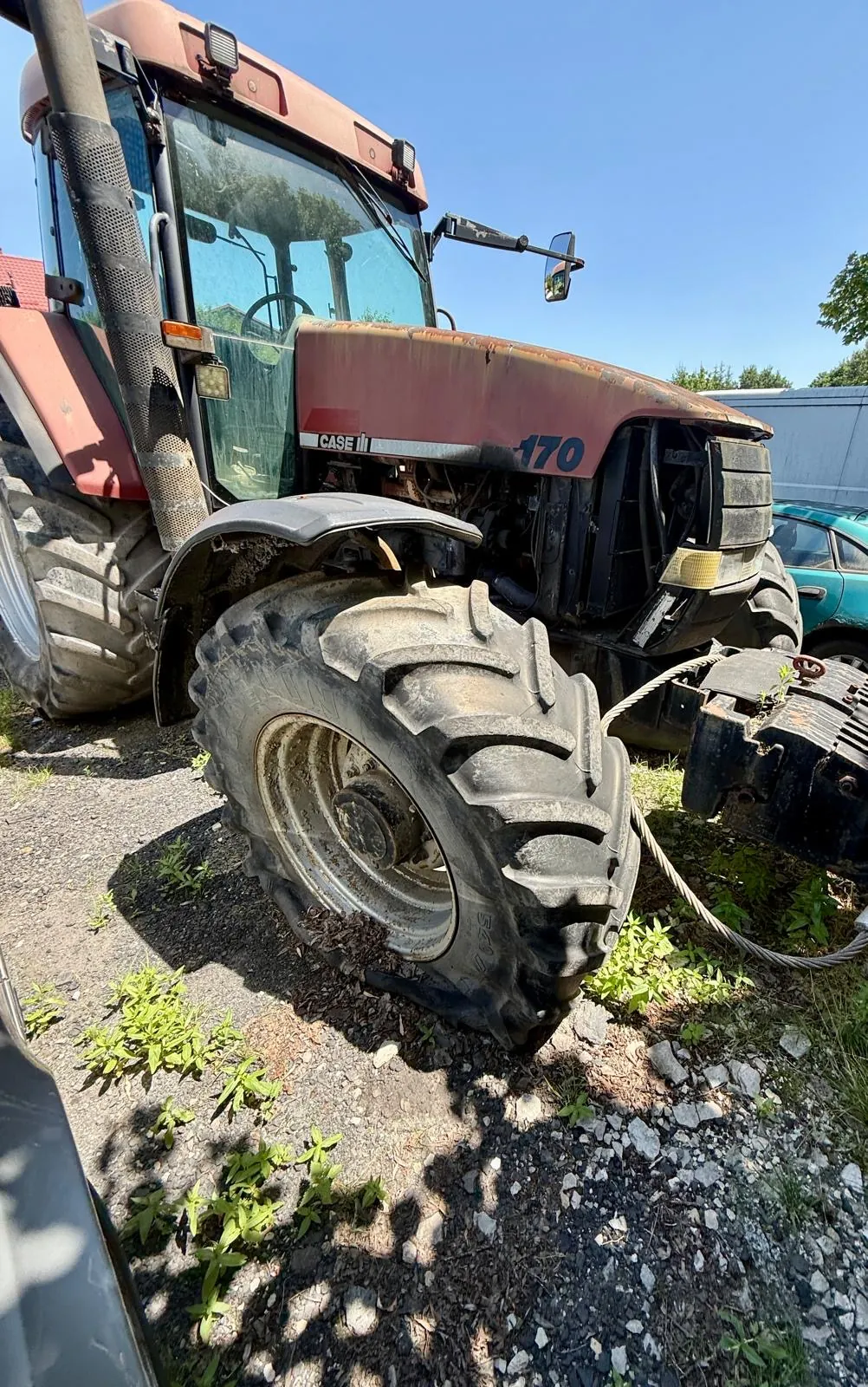 Zbliżenie na przednie lewe koło dużego, czerwonego traktora marki Case IH z numerem 170 na masce. Traktor stoi na szutrowym podłożu z kępami zielonej trawy. Widoczna jest kabina kierowcy z szybami oraz fragmenty mechanizmów maszyny.