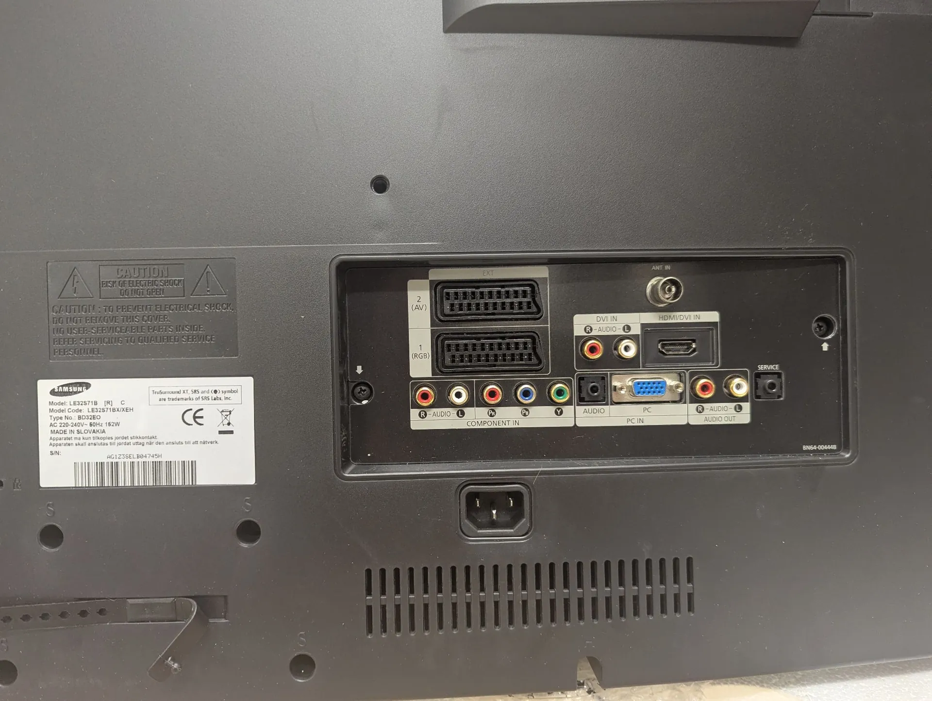 Tylny panel monitora lub telewizora z zestawem portów wejściowych i wyjściowych. Widoczne są dwa gniazda VGA (oznaczone jako 2 (AV) i 1 (RGB)), gniazda RCA (żółte, białe, czerwone), gniazdo DVI-IN, HDMI/DVI-IN oraz gniazdo zasilania AC. Po lewej stronie znajduje się naklejka z ostrzeżeniami w języku angielskim oraz tabliczka znamionowa z danymi technicznymi: Model: LS27T571B, Model Code: LS27T571B/XEH, Typ: Monitor, AC 220-240V~ 50Hz - 180W, Made in Slovakia. Na dole widoczna jest kratka wentylacyjna.