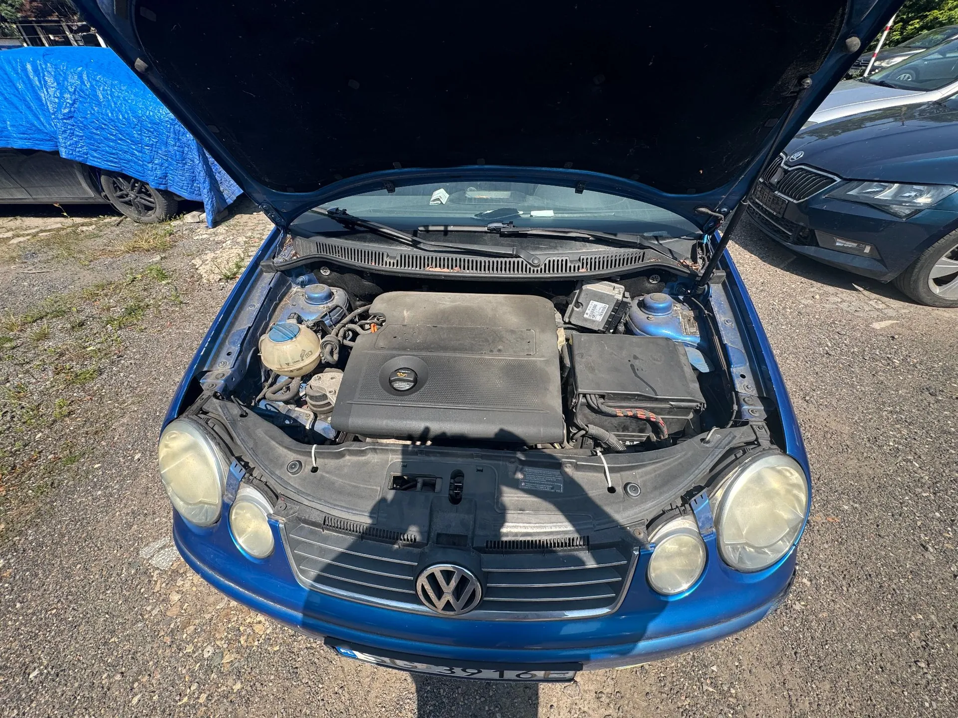 Niebieski samochód marki Volkswagen z otwartą maską, ukazującą komorę silnika. Widoczna jest czarna osłona silnika z logo VW, zbiornik płynu chłodniczego oraz akumulator. W tle widoczne są inne samochody, w tym jeden przykryty niebieską plandeką.