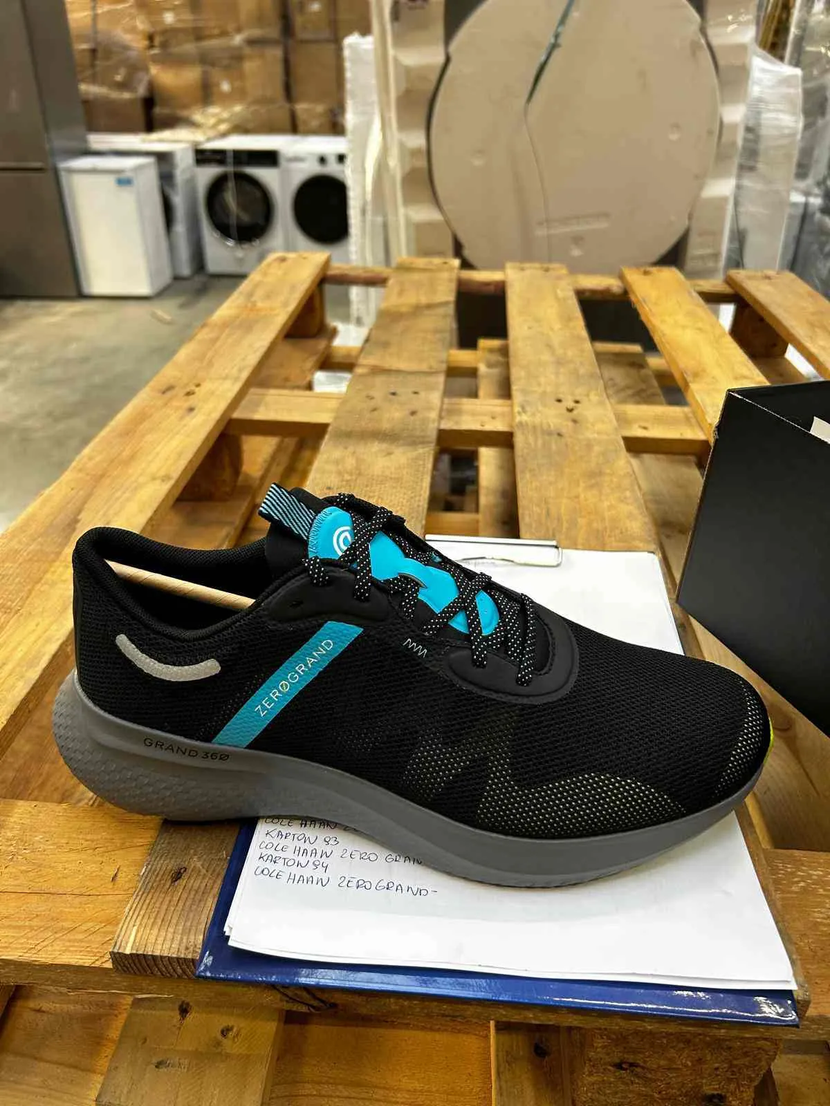 Czarny but sportowy z niebieskimi akcentami i szarą podeszwą, umieszczony na drewnianym palecie. Na boku buta widoczny napis 'ZEROGRAND' oraz 'GRAND 360'. But leży na białym papierze z ręcznie zapisanymi notatkami: 'KARTON 93', 'COLE HAAN ZERO GRAND', 'KARTON 94', 'COLE HAAN ZERO GRAND-'. W tle białe pralki, pudełka i okrągły element. Obok buta czarna pudełko i niebieski schowek.