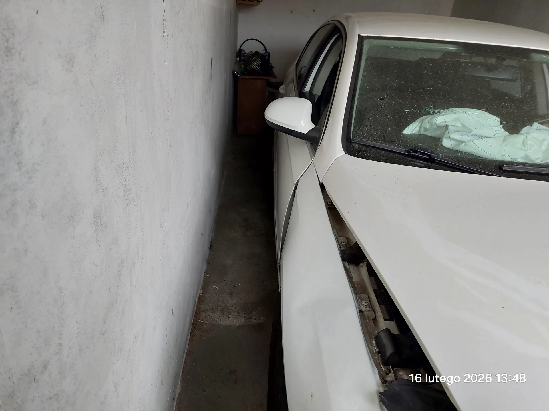 Biały samochód zaparkowany w wąskim przejściu między białą ścianą a inną konstrukcją. Kapota samochodu jest otwarta, odsłaniając części silnika. Na przedniej szybie leży jasnoniebieska tkanina. W tle widoczna jest drewniana szafka z koszem na górze. Podłoga z betonu, ściany są białe i matowe. W prawym dolnym rogu obrazu znajduje się znacznik czasu: 16 lutego 2026 13:48.
