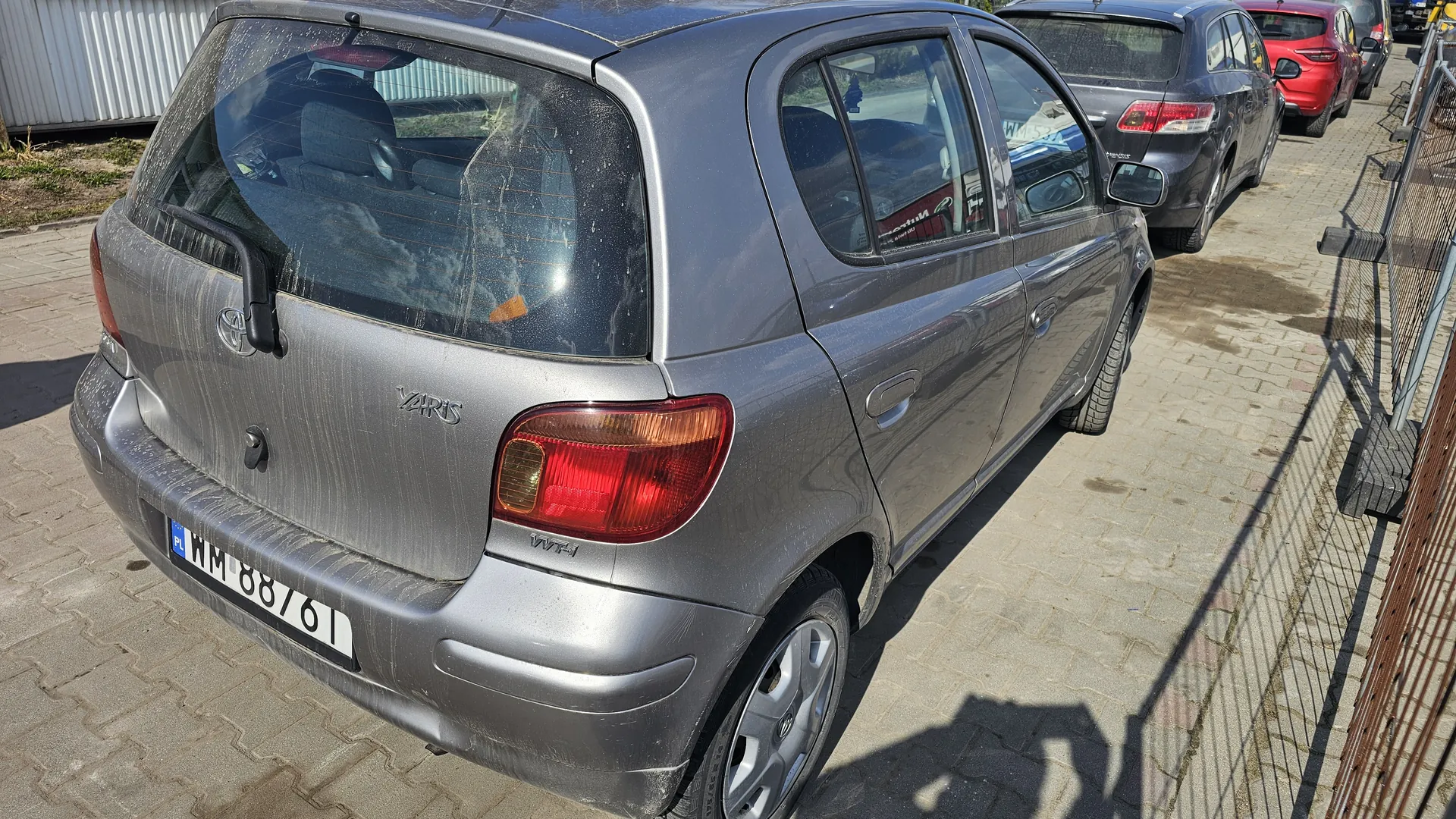 Srebrny samochód Toyota Yaris zaparkowany na brukowanej powierzchni. Widok z tyłu i boku. Na tylnej szybie widoczne ślady brudu, a na bagażniku napis "YARIS" oraz emblemat marki. Tablica rejestracyjna z białym tłem i czarnymi literami/ cyframi: WM 88761, z niebieskim pasem po lewej stronie z napisem "PL". Na prawo od samochodu metalowe ogrodzenie, w tle inne zaparkowane pojazdy (szary i czerwony) oraz budynek z białymi blachami. Słoneczny dzień, cienie widoczne na ziemi.