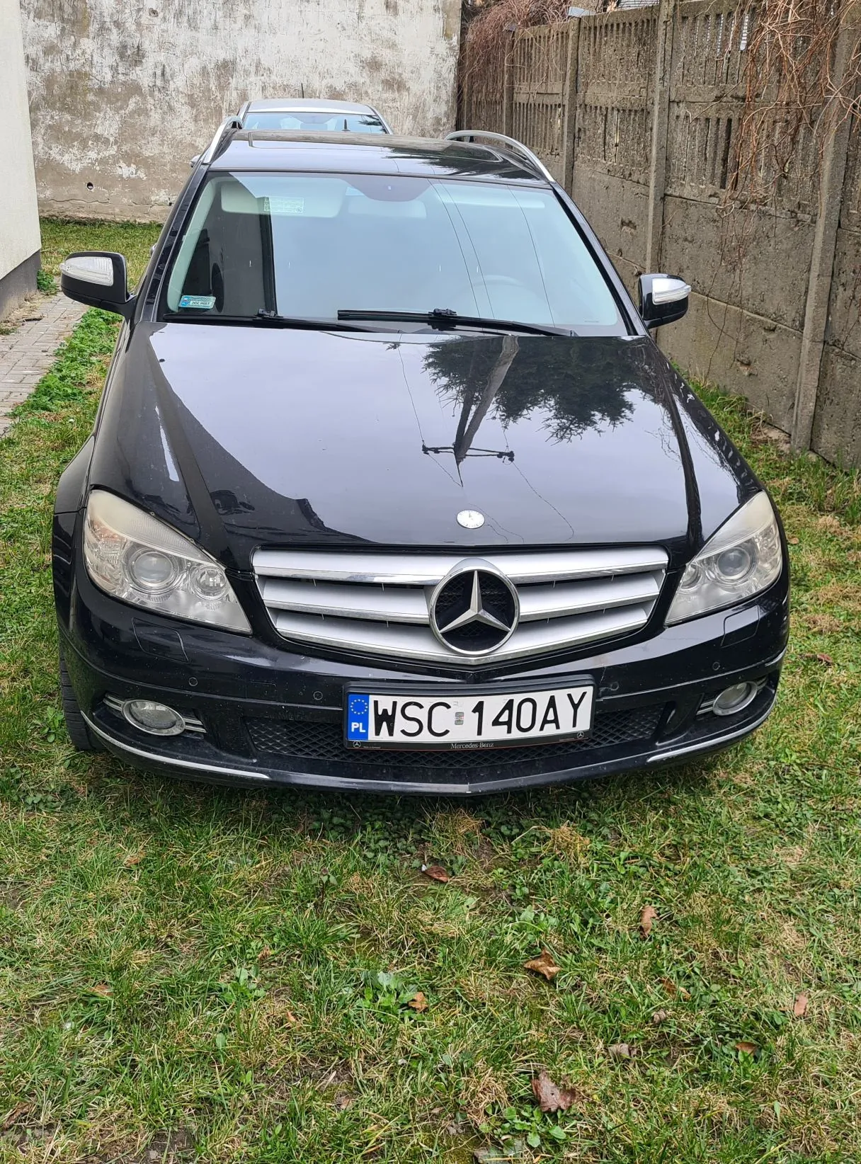 Czarny samochód marki Mercedes-Benz zaparkowany na trawie. Widoczna tablica rejestracyjna z napisem PL WSC 140AY. W tle betonowa ściana, ogrodzenie i trawa. Samochód ma przednie światła, chromowaną atrapę chłodnicy z logo trójgwiazdy oraz dach z przenośnymi bagażnikami. Na przedniej szybie znajduje się niebieski znaczek.