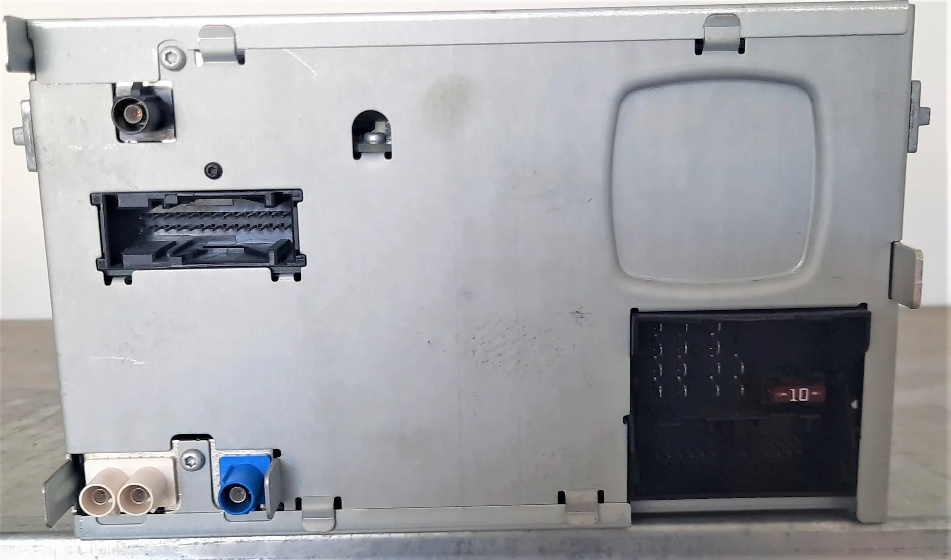Zdjęcie przedstawiające tylną, metalową obudowę urządzenia elektronicznego, prawdopodobnie radia samochodowego. Widoczne są różne złącza: w lewym górnym rogu gniazdo antenowe, poniżej szerokie czarne złącze wielopinowe, w prawym dolnym rogu duże czarne gniazdo z napisem '-10-', a w lewym dolnym rogu dwa białe gniazda RCA oraz jedno niebieskie gniazdo RCA.