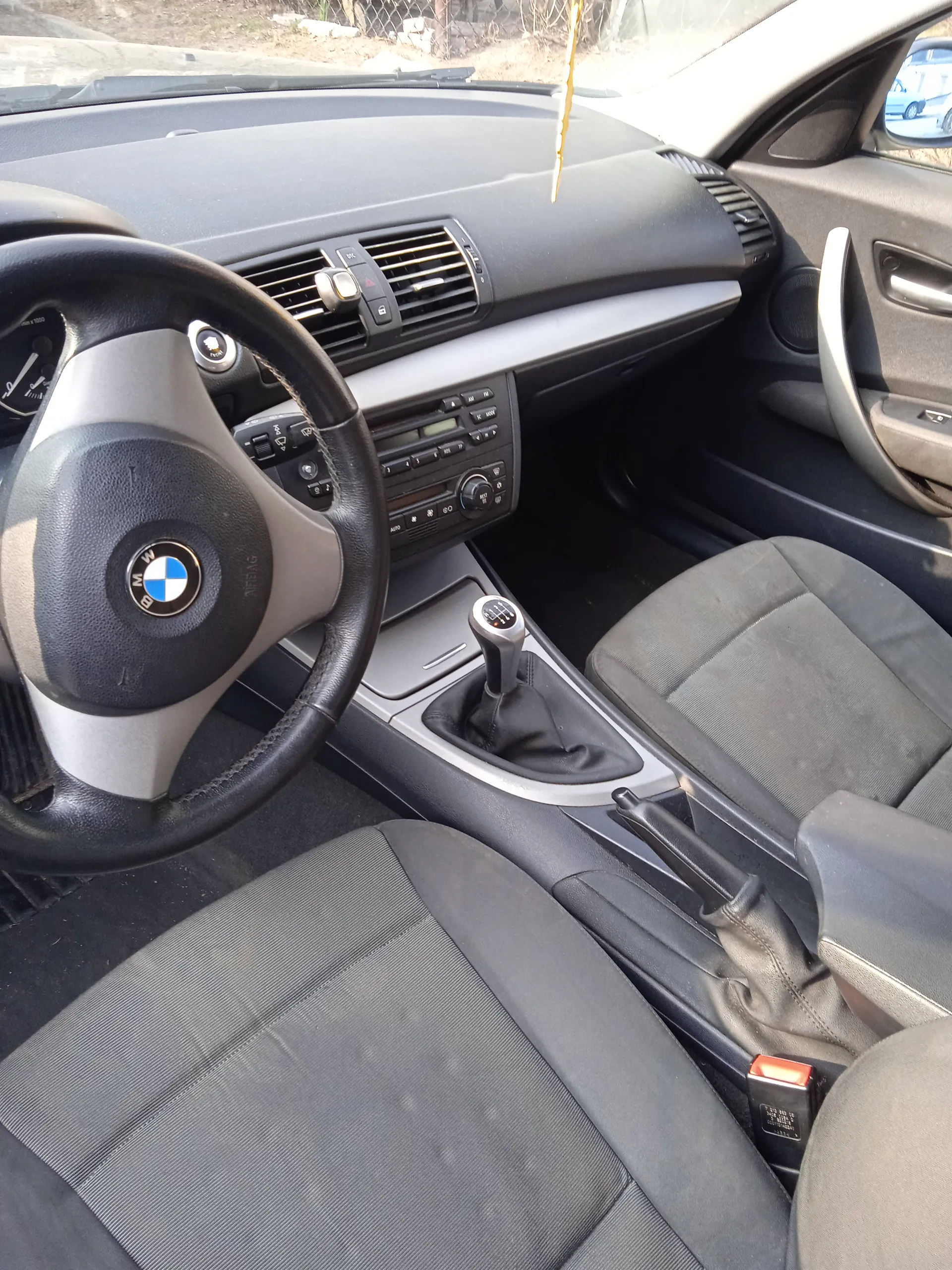 Wnętrze samochodu marki BMW z widocznym kierownicą z logo, deską rozdzielczą, przyciskami klimatyzacji, radio, dźwignią zmiany biegów i siedzeniami w kolorze szarym. W tle przez przednią szybę widać ogrodzenie i fragment innego pojazdu. Na lusterku wstecznym wiszący przedmiot.