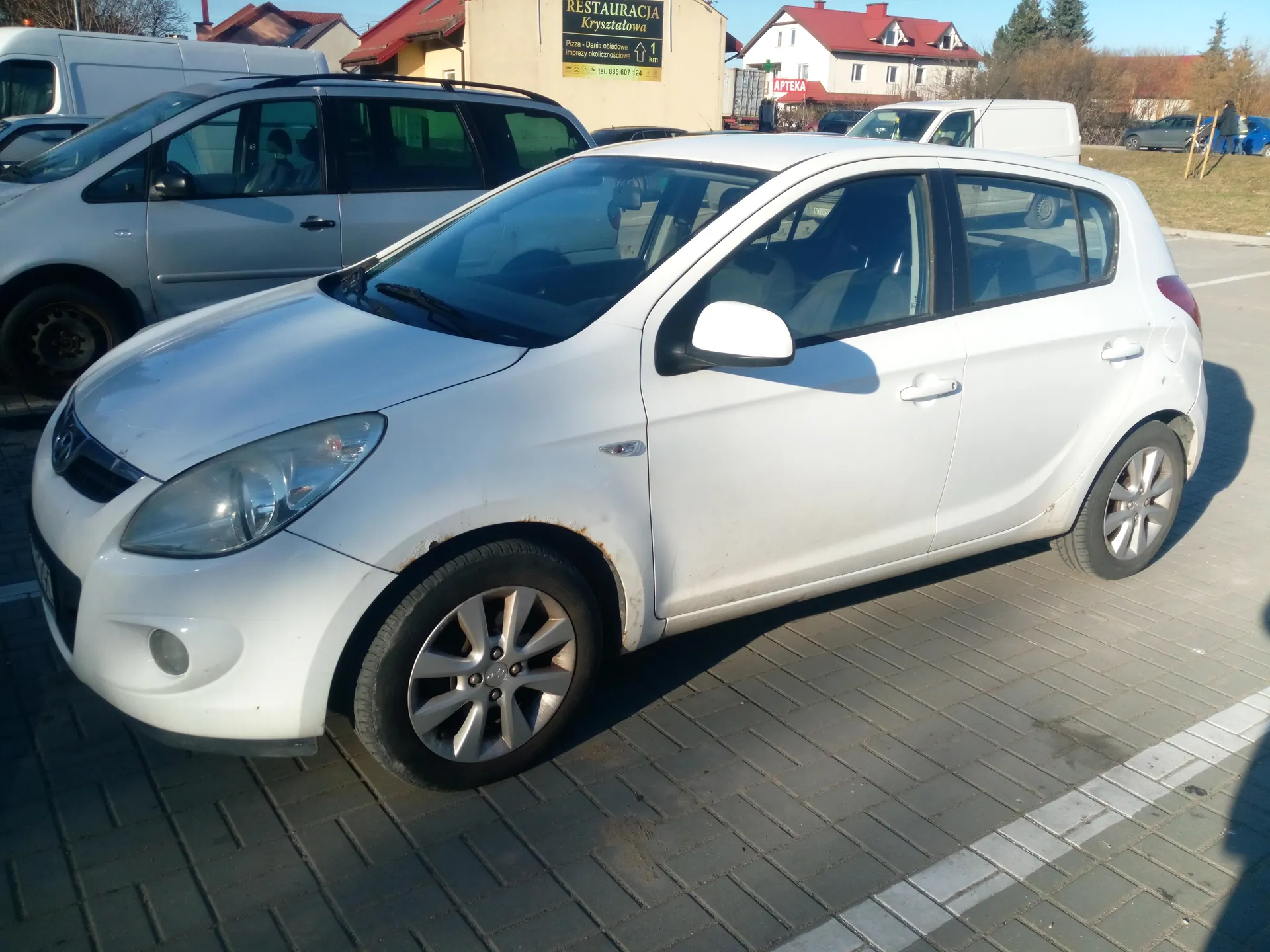 Biały samochód osobowy typu hatchback zaparkowany na brukowanym placu parkingowym. W tle widoczne są inne pojazdy: srebrny minivan i białe furgony. Za nimi budynki z widocznymi szyldami: 'RESTAURACJA Kryształowa' z dodatkowym napisem 'Pizzeria - Dania obiadowe imprezy okolicznościowe' oraz 'APTEKA'. Samochód ma srebrne felgi, widoczne ślady rdzy w okolicach błotników. Na tle drzewa bez liści i jasne niebo. Tekst na szyldach jest czytelny i poziomo ustawiony.