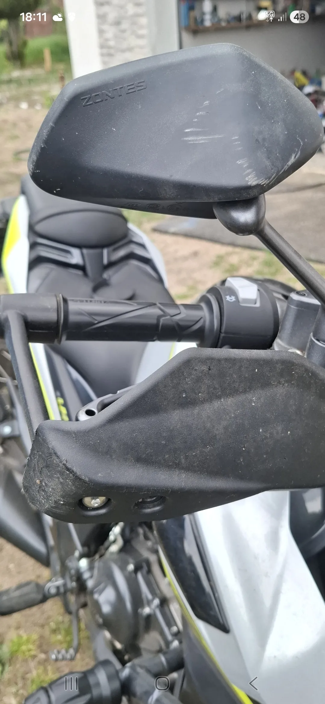 Zbliżenie na czarne lusterka boczne motocykla z widocznym napisem ZONTES na górnym lusterku. Widoczne są czarne kierownice z przyciskami, czarno-żółty motocykl oraz tło z trawą, betonową powierzchnią i fragmentem budynku. Na górze zdjęcia widoczny jest pasek stanu telefonu z godziną 18:11, ikonami sieci 5G i poziomem baterii 48%.