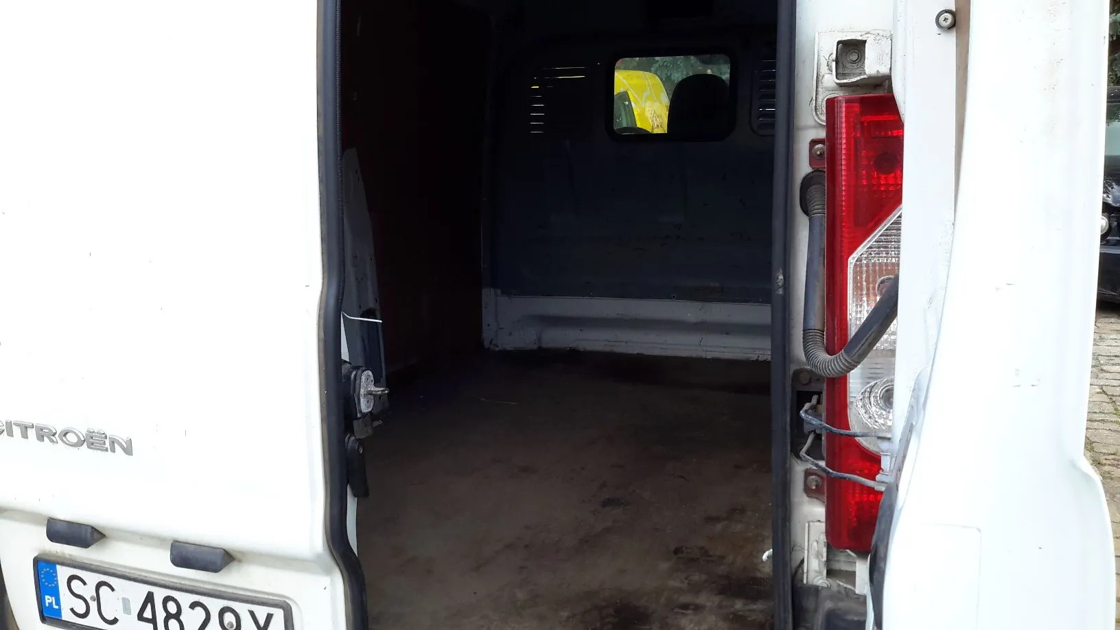 Widok z tyłu białego samochodu dostawczego z otwartymi tylnymi drzwiami, ukazujący pustą przestrzeń ładunkową. Po lewej stronie widoczny jest fragment napisu 'CITROËN' oraz tablica rejestracyjna o numerze 'SC 4820V' z oznaczeniem 'PL'. Po prawej stronie znajduje się pionowa lampa tylna w kolorze czerwonym.