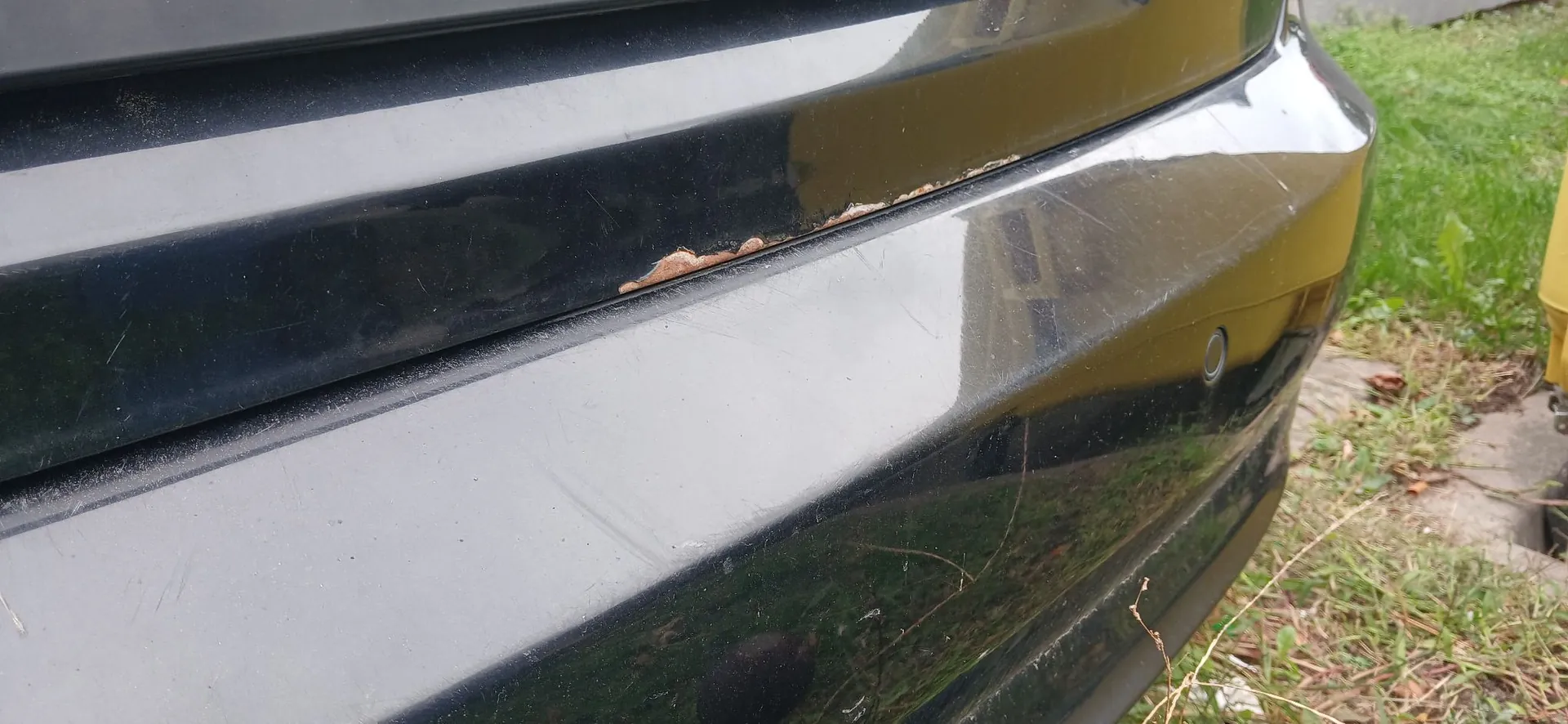 Zbliżenie na czarny, lakierowany element nadwozia samochodu, prawdopodobnie zderzak. Na krawędzi między dwiema częściami elementu widoczne jest odpryśnięcie lakieru i rdza. Powierzchnia lakieru posiada liczne drobne zarysowania.