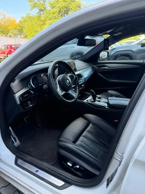 Widok z otwartych drzwi kierowcy do wnętrza białego samochodu marki BMW. Widoczna jest czarna skórzana tapicerka foteli, czarna kierownica z logo BMW w centrum, deska rozdzielcza z ekranem multimedialnym oraz konsola środkowa z lewarkiem zmiany biegów. Na progu samochodu znajduje się srebrna listwa z kolorowym logo M.