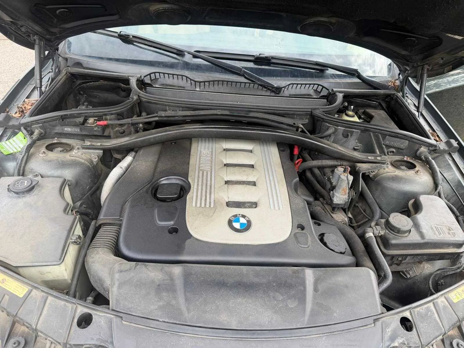 Otwarty kapota samochodu marki BMW z widocznym silnikiem. Na pokrywie silnika znajduje się logo BMW oraz napis 'BMW'. Wokół widoczne są różne elementy silnika, w tym zbiorniki, przewody i wycieraczki przednie. Kapota jest podparta, a w tle widać przednią szybę.