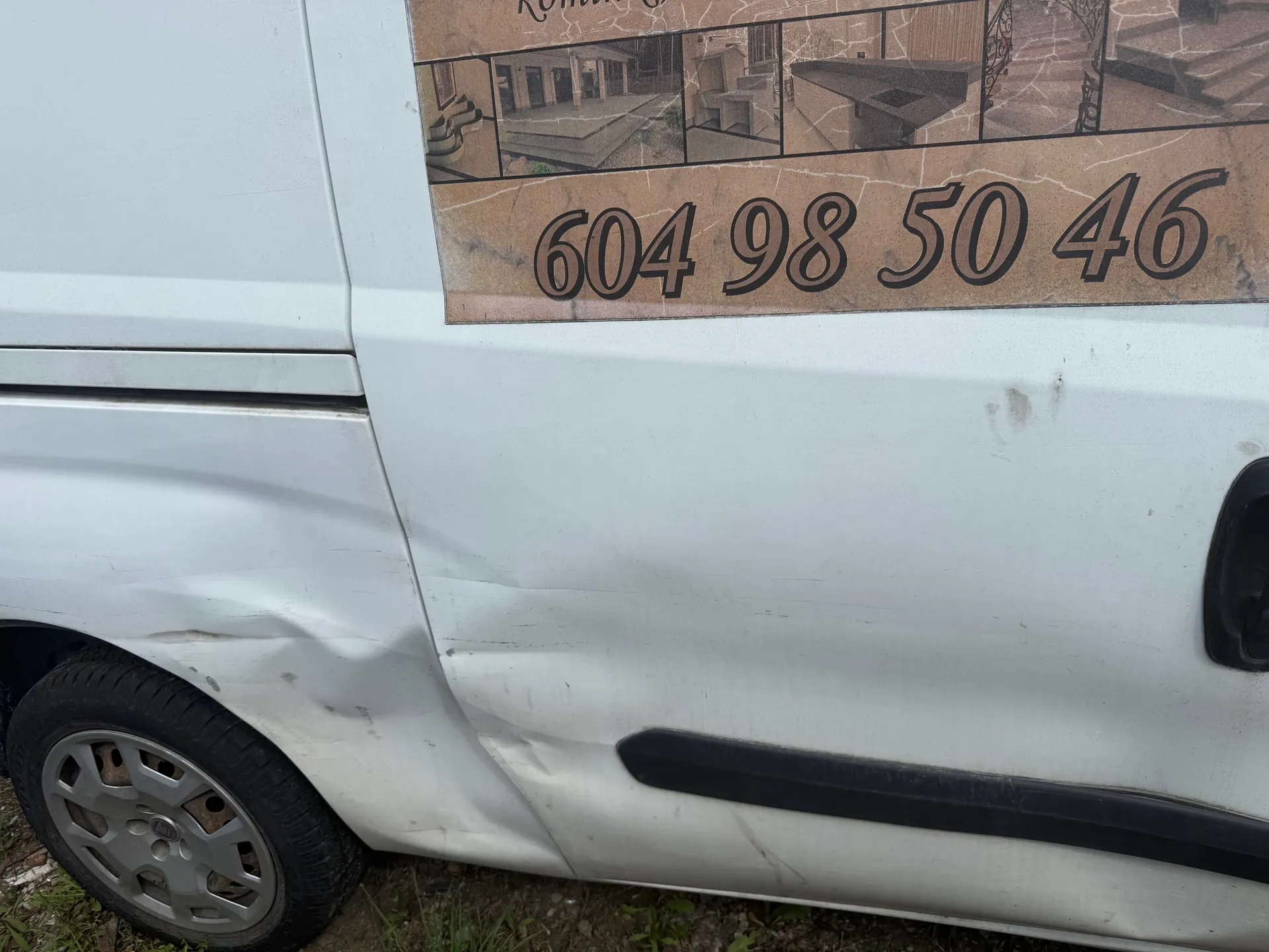 Biały samochód dostawczy z widocznym wgnieceniem na boku, kołem z aluminiową felgą i czarną obwódką. Na boku pojazdu naklejka z brązowym tłem, na której znajdują się zdjęcia budynków i schodów oraz numer telefonu 604 98 50 46. Samochód stoi na trawie i ziemi.