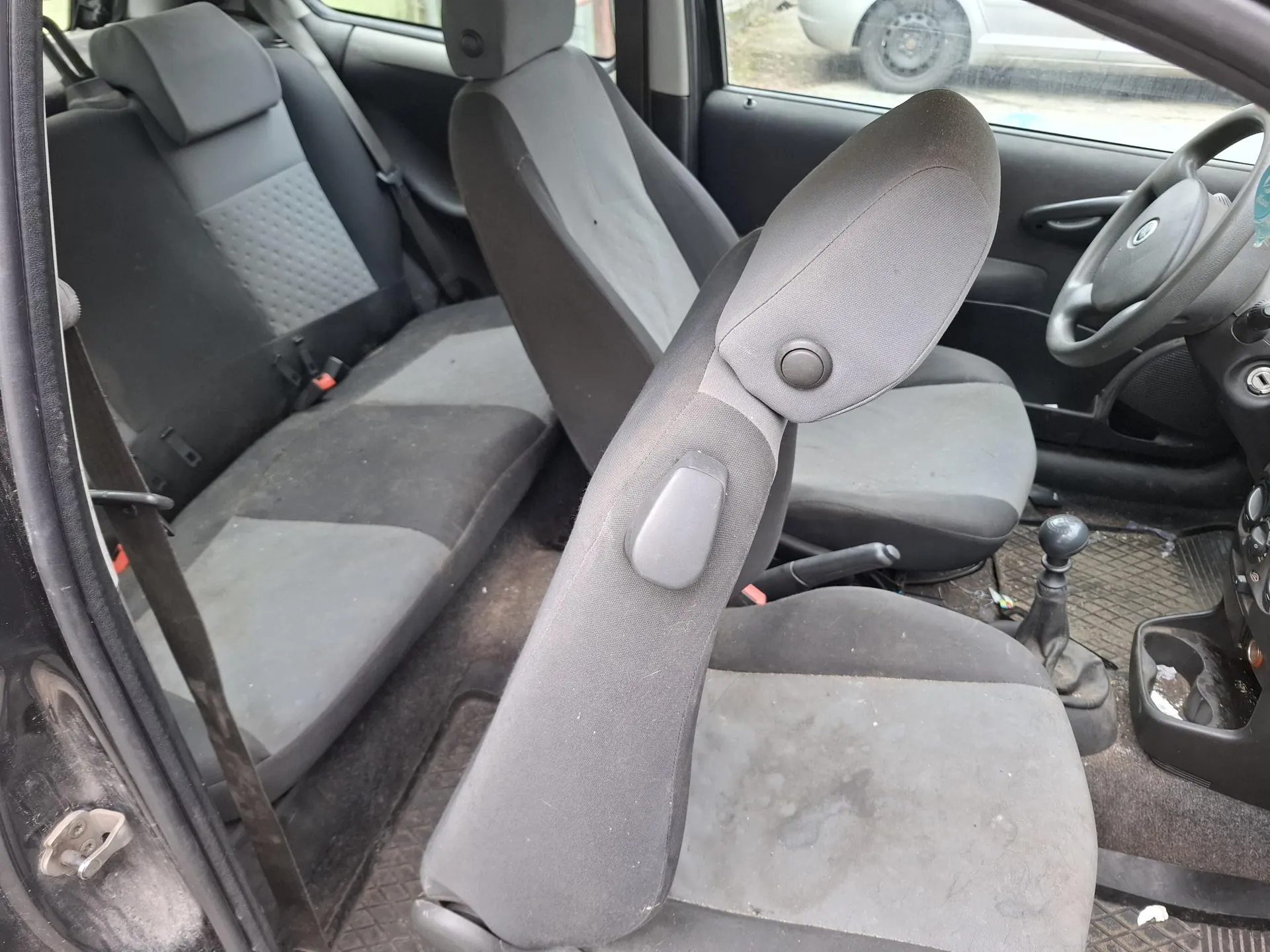 Wnętrze samochodu z szarymi tkaninowymi fotelami, fotel kierowcy z podłokietnikiem i zagłówkiem, kierownicą po prawej stronie, dźwignią zmiany biegów oraz otwartą drzwiami. Widoczne są ślady brudu na siedzeniach i podłodze. Poza samochodem częściowo widoczny inny pojazd.