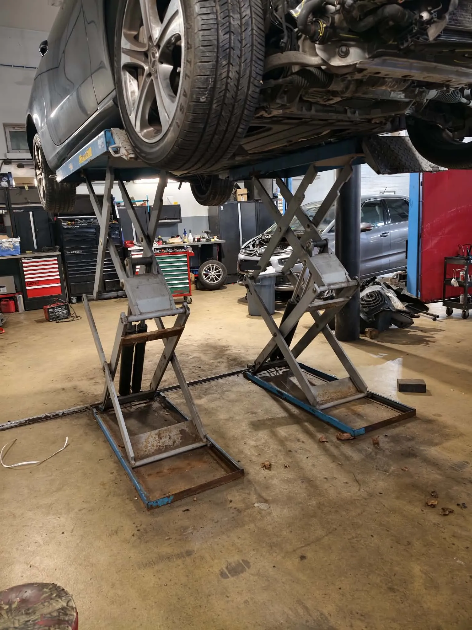 Samochód osobowy w kolorze ciemnoszarym uniesiony na hydraulicznym podnośniku nożycowym w warsztacie samochodowym. Widoczny jest fragment przedniego koła z felgą aluminiową oraz podwozie pojazdu. W tle znajdują się szafki narzędziowe, inny samochód oraz wyposażenie warsztatu.