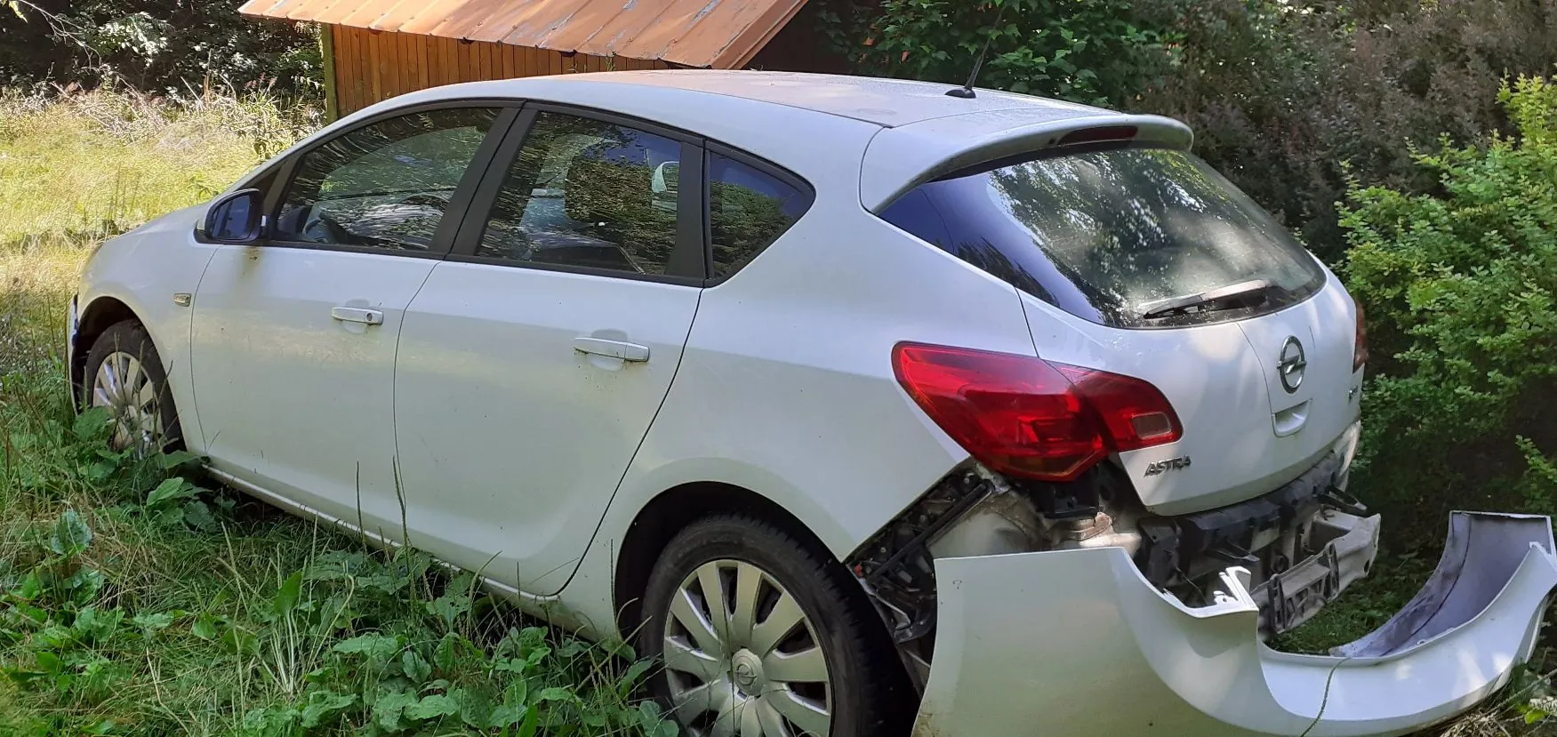 Biały samochód marki Opel Astra zaparkowany w wysokiej trawie. Pojazd jest uszkodzony z tyłu – tylny zderzak jest częściowo oderwany i zwisa ku dołowi, odsłaniając wewnętrzne elementy konstrukcyjne. W tle widoczny jest fragment drewnianego budynku oraz zielone krzewy i drzewa.