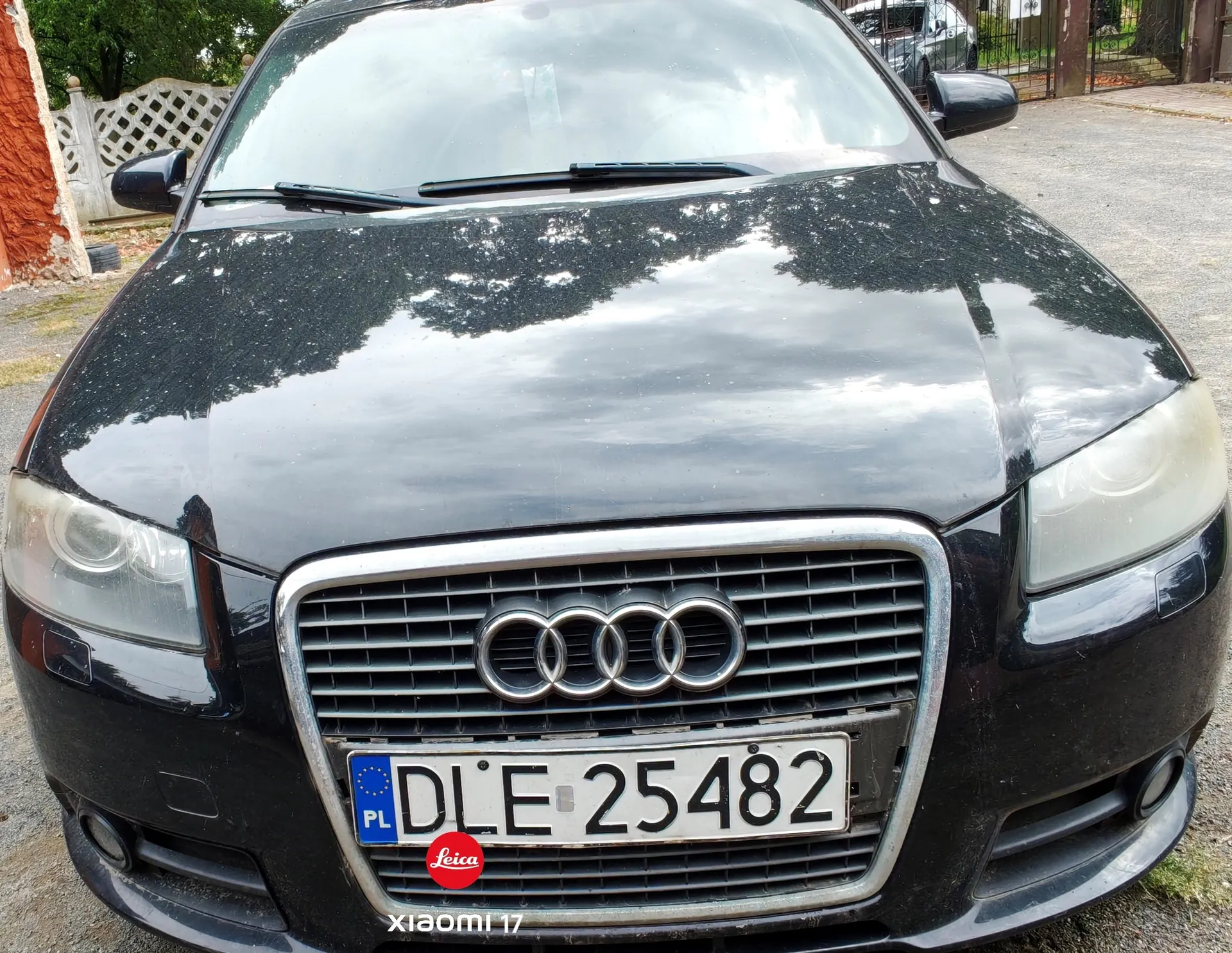 Widok z przodu na czarny samochód marki Audi. Widoczna jest maska, przednie reflektory oraz charakterystyczny grill z logo czterech pierścieni. Samochód posiada polską tablicę rejestracyjną o numerze DLE 25482, pod którą znajduje się mała czerwona naklejka z napisem 'Leica'. Na dole obrazu widnieje biały napis 'xiaomi 11T'.