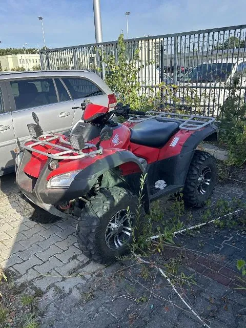Czerwony quad marki Honda zaparkowany na kostce brukowej obok srebrnego samochodu typu kombi. Quad posiada przedni i tylny bagażnik, duże terenowe opony oraz widoczne logo Hondy na boku. W tle znajduje się metalowy płot oraz inne zaparkowane samochody.