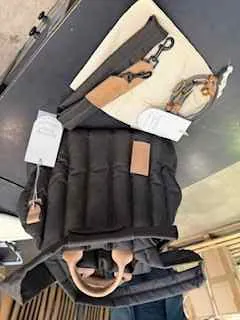 Czarna pikowana kurtka z brązową skórzaną naszywką na klatce piersiowej i białą etykietą. Obok kurtki znajduje się beżowa torba na ramię z brązowym paskiem i metalowym zamkiem oraz brązowy spleciony akcesorium. W tle ciemna powierzchnia stołu i kartonowe pudełka. Tekst na etykietach nieczytelny.