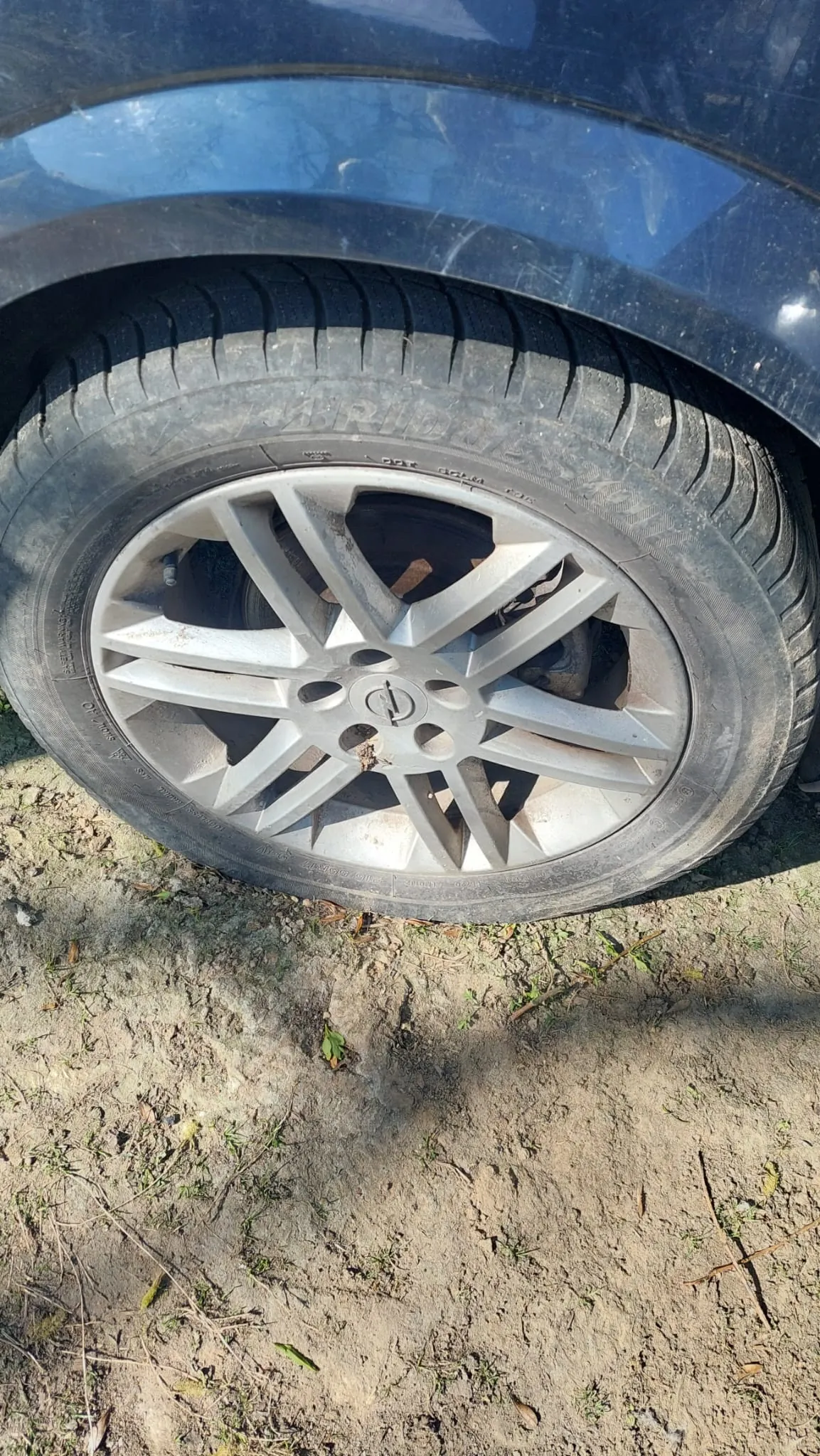 Zbliżenie na koło samochodu z oponą marki BRIDGESTONE. Widoczny srebrny felga z pięcioma ramionami, czarna opona z widocznym profilem i fragmentem ciemnoniebieskiego nadwozia. Pod kołem brudna ziemia z rzadką trawą i suchymi gałązkami.
