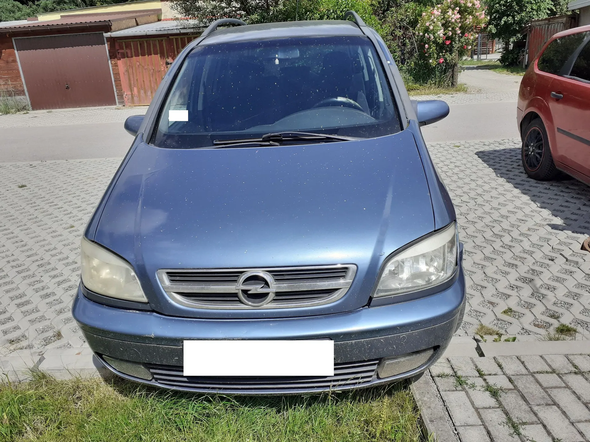 Widok z przodu niebieskiego samochodu marki Opel zaparkowanego na kostce brukowej. Pojazd ma srebrny grill z logo producenta w centrum oraz białą, zakrytą tablicę rejestracyjną. W tle widoczne są garaże, zieleń oraz fragment czerwonego samochodu po prawej stronie.