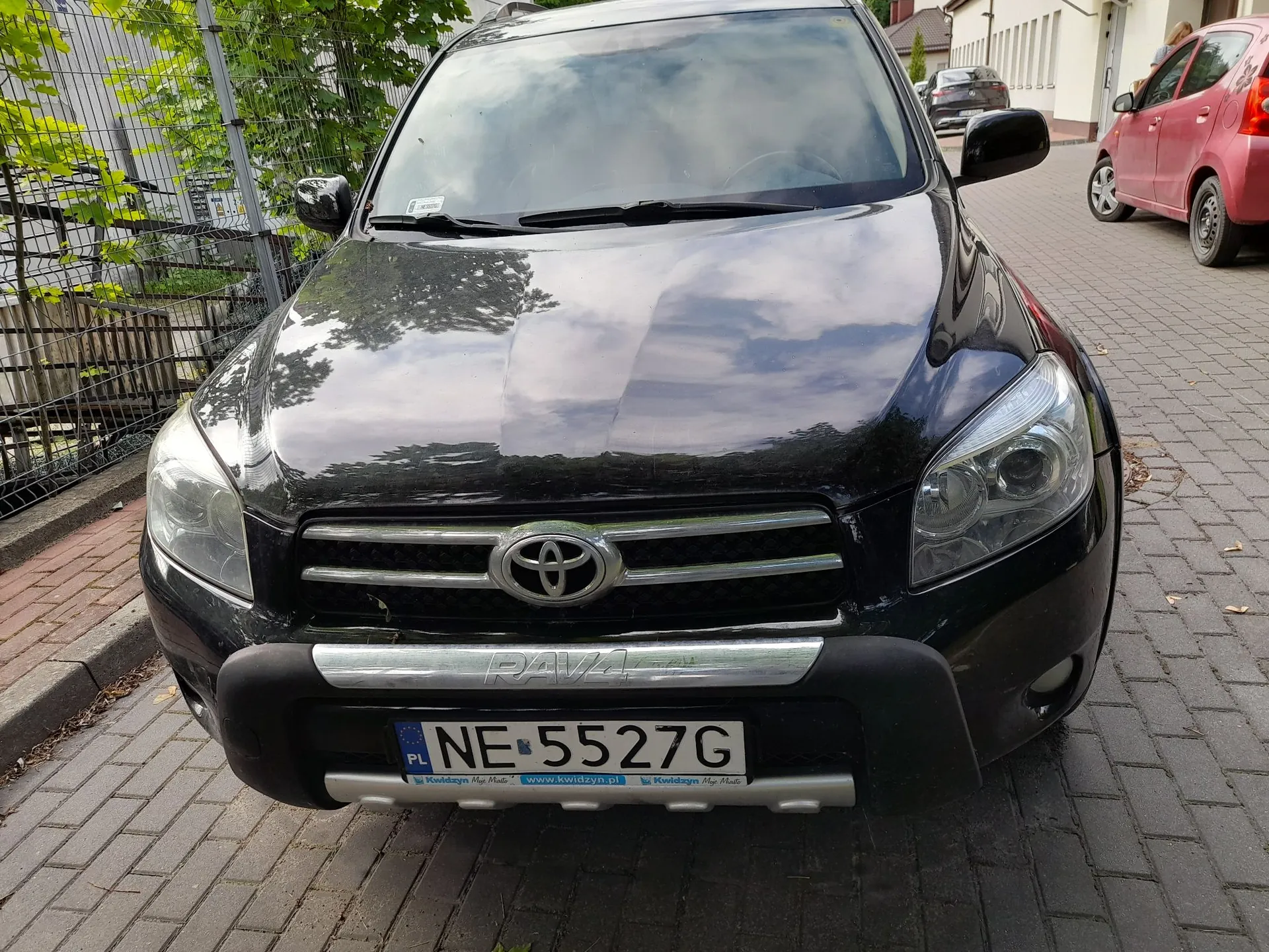 Czarny samochód marki Toyota RAV4 zaparkowany na kostce brukowej. Widoczny jest przód pojazdu z grillem, logo Toyoty oraz tablicą rejestracyjną o numerze NE 5527G. Poniżej numeru rejestracyjnego znajduje się napis 'Kwidzyn Moje Miasto' oraz adres strony internetowej 'www.kwidzyn.pl'. W tle widoczne są zielone krzewy, ogrodzenie oraz fragment czerwonego samochodu po prawej stronie.