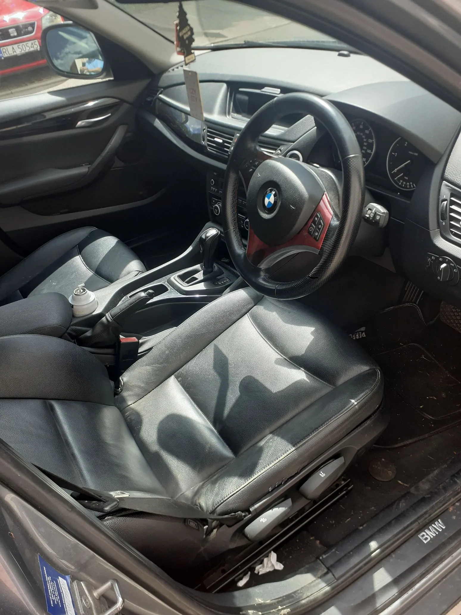 Wnętrze samochodu marki BMW z perspektywy otwartych drzwi kierowcy. Widoczny jest czarny skórzany fotel, kierownica z logo BMW, deska rozdzielcza, konsola środkowa z lewarkiem zmiany biegów oraz fragmenty drzwi. Przez przednią szybę widoczny jest fragment innego czerwonego samochodu z tablicą rejestracyjną RL A 50545.