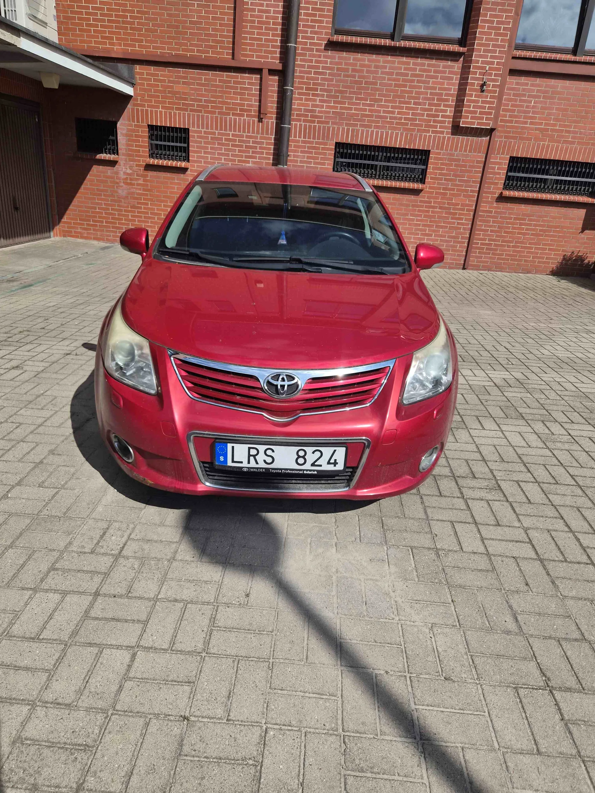 Czerwony samochód marki Toyota zaparkowany na kostce brukowej przed budynkiem z czerwonej cegły. Widoczna jest tablica rejestracyjna o numerze LRS 824.