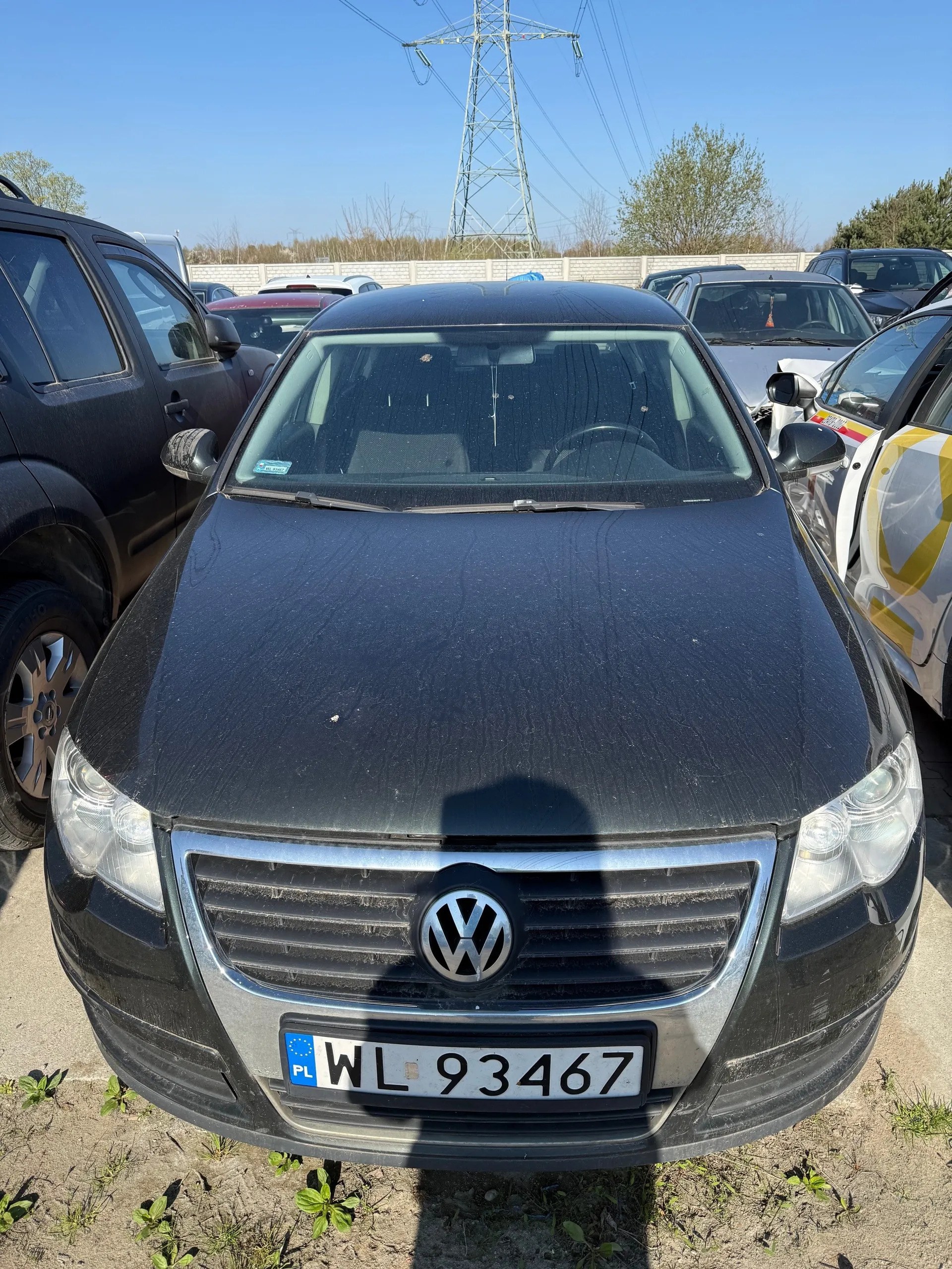 Czarny samochód marki Volkswagen z tablicą rejestracyjną PL WL 93467, zaparkowany na otwartym placu. Na pierwszym planie widoczna przód pojazdu z logo VW, zanieczyszczonym nadwoziem i reflektorami. W tle inne zaparkowane samochody, betonowy mur, drzewa, wysoka wieża energetyczna oraz jasne niebo. Na ziemi rosną pojedyncze rośliny.