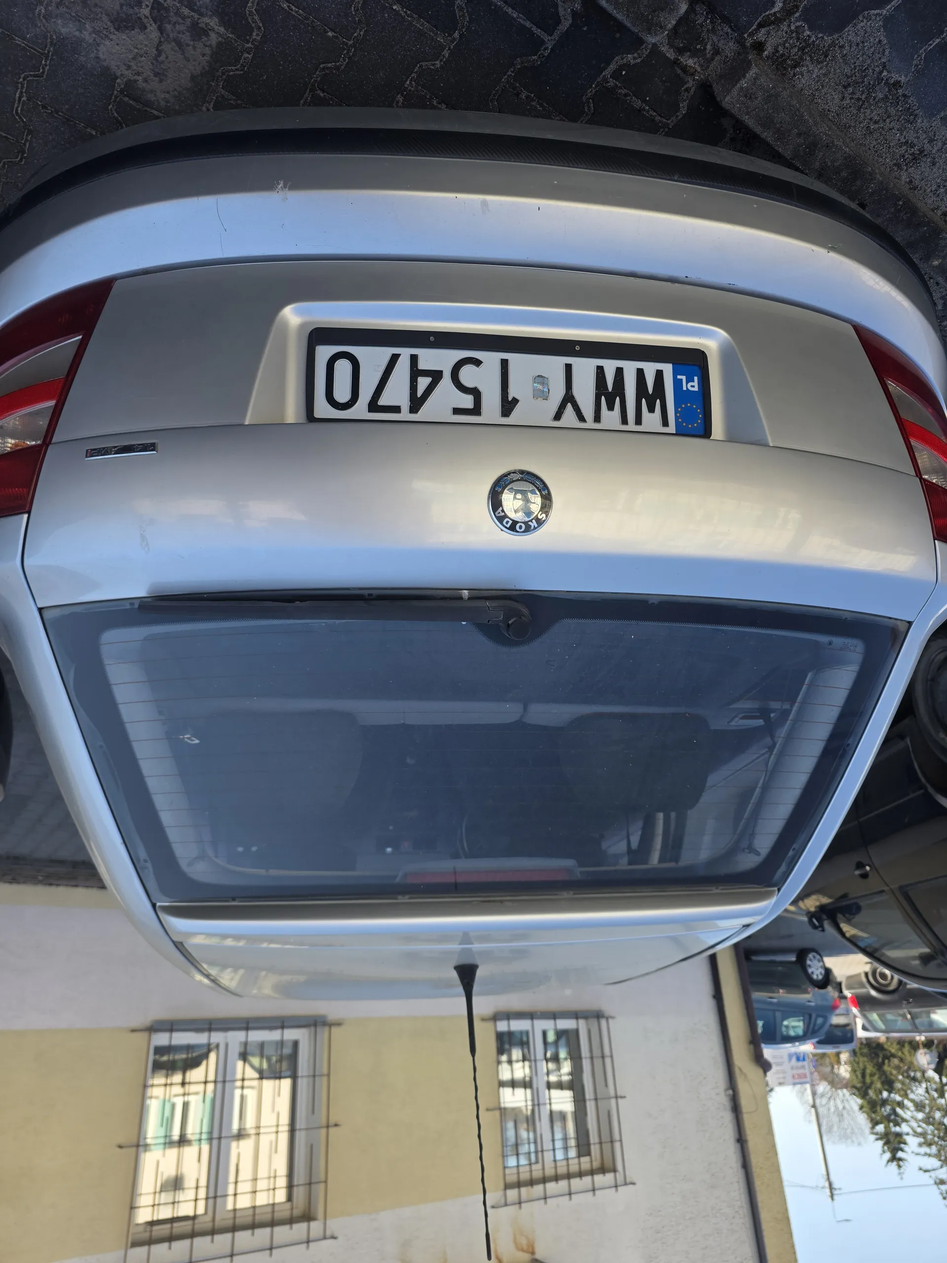Z tyłu srebrny samochód marki Skoda z tablicą rejestracyjną WWY 15470. Tablica posiada niebieski pas z napisem PL i flagą Unii Europejskiej. W tle budynek z oknami osłoniętymi kratą oraz inne zaparkowane pojazdy na brukowanej nawierzchni.