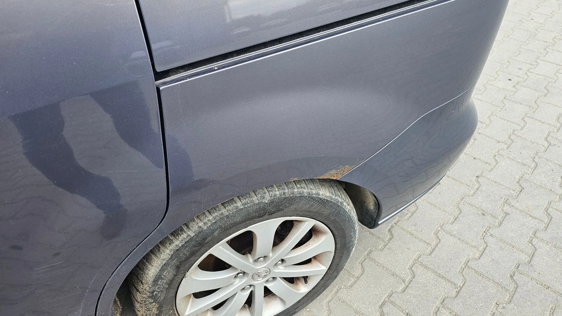 Zbliżenie na tylny prawy błotnik i koło samochodu w kolorze ciemnofioletowym. Na krawędzi nadkola widoczne jest niewielkie ognisko korozji (rdza). Koło posiada srebrną felgę aluminiową z pięcioma podwójnymi ramionami.