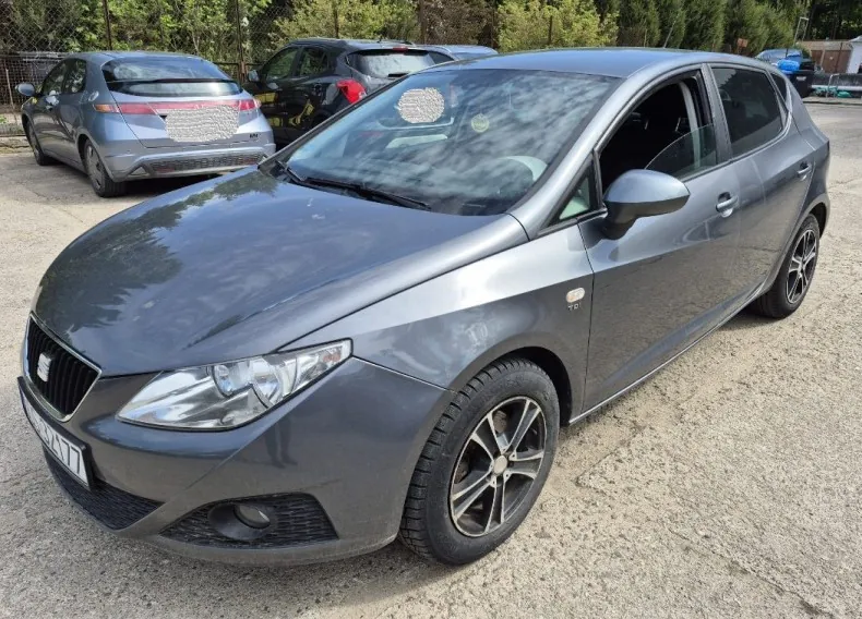 Szary samochód marki SEAT (model Leon) zaparkowany na żwirowym placu. Widoczna przód pojazdu z logo marki, reflektory i częściowo widoczna tablica rejestracyjna WU4477. W tle inne samochody (srebrny i czarny), ogrodzenie i drzewa. Powierzchnia podłogi pokryta żwirem, a na tle zielone krzewy i drzewa.