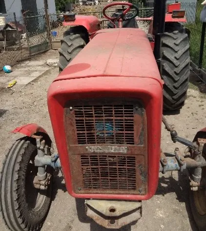 Czerwony traktor rolniczy zaparkowany na piaszczystym podłożu. Widoczny jest przód maszyny z metalową kratką chłodnicy, przednie koła oraz tylne koła z charakterystycznym bieżnikiem. W tle widać ogrodzenie z siatki oraz fragmenty budynków.